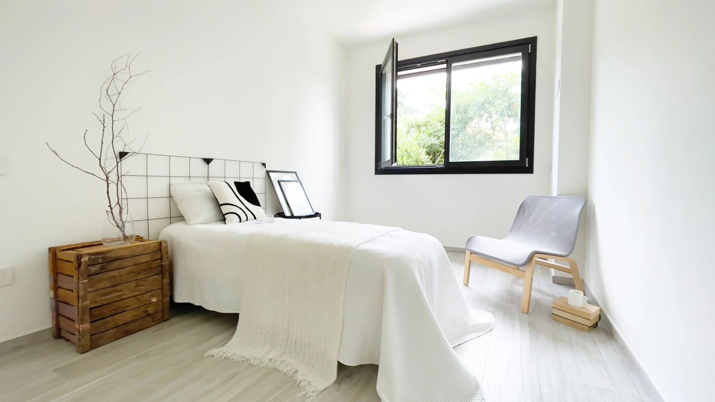 Camera da letto minimalista con letto bianco, cuscini decorativi, sedia moderna, finestra aperta, pavimento in legno chiaro e tavolino con vaso di rami secchi.