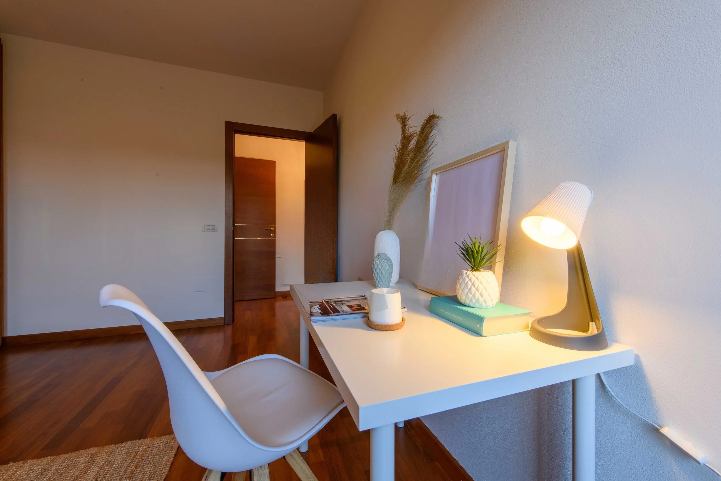Scrivania bianca con lampada, vaso di piante, libri e sedia moderna in una stanza con pavimento in legno e porta aperta.