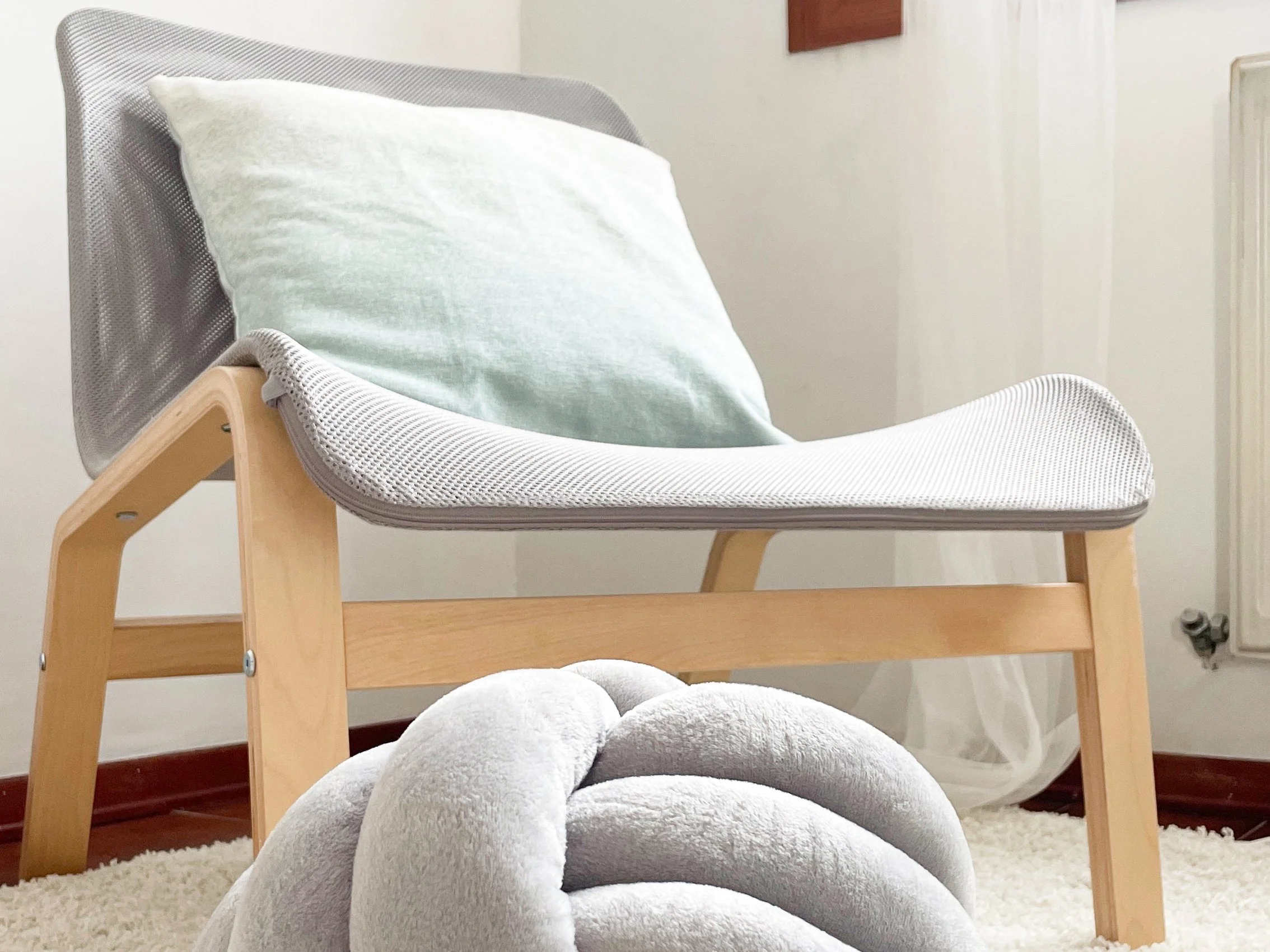 Sedia moderna con struttura in legno chiaro e seduta in tessuto grigio, con un cuscino verde chiaro. Davanti alla sedia, un cuscino decorativo nodo grigio chiaro su tappeto bianco.