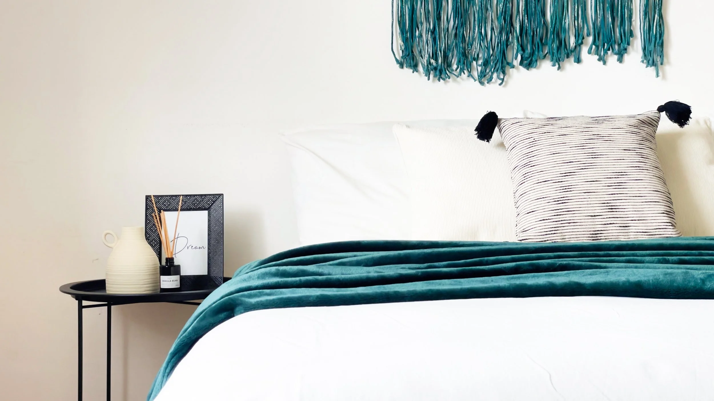 Camera da letto con un letto bianco, coperta verde, cuscini decorativi e una mensola con un vaso e diffusore di profumo.