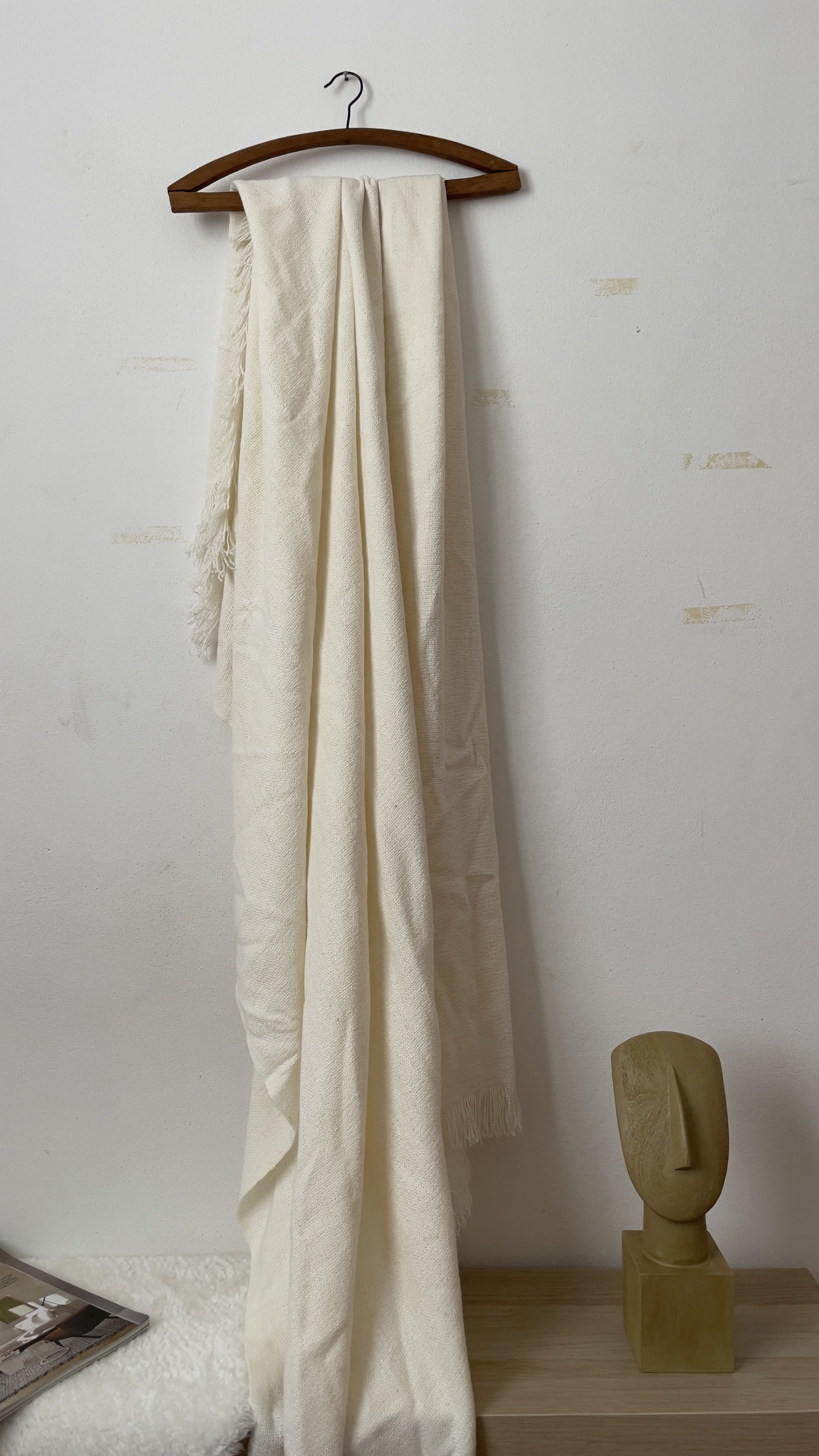Una sciarpa bianca appesa a una gruccia di legno sopra un tavolo, accanto a una scultura astratta di legno e una rivista.