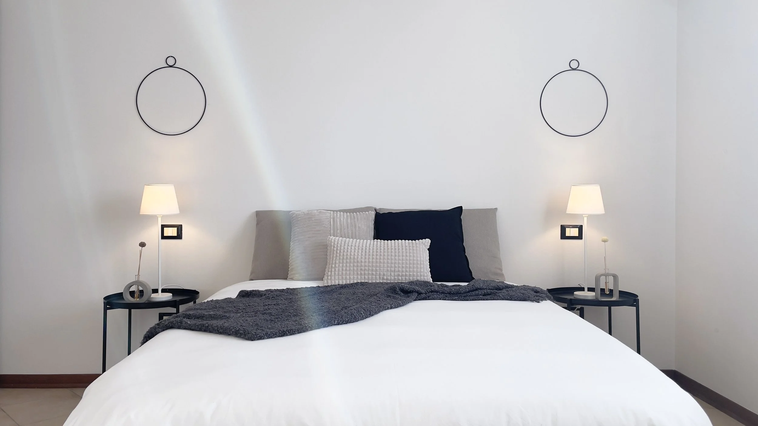 Camera da letto moderna con letto matrimoniale, cuscini decorativi, coperte, due comodini con lampade e decorazioni minimaliste sulla parete.
