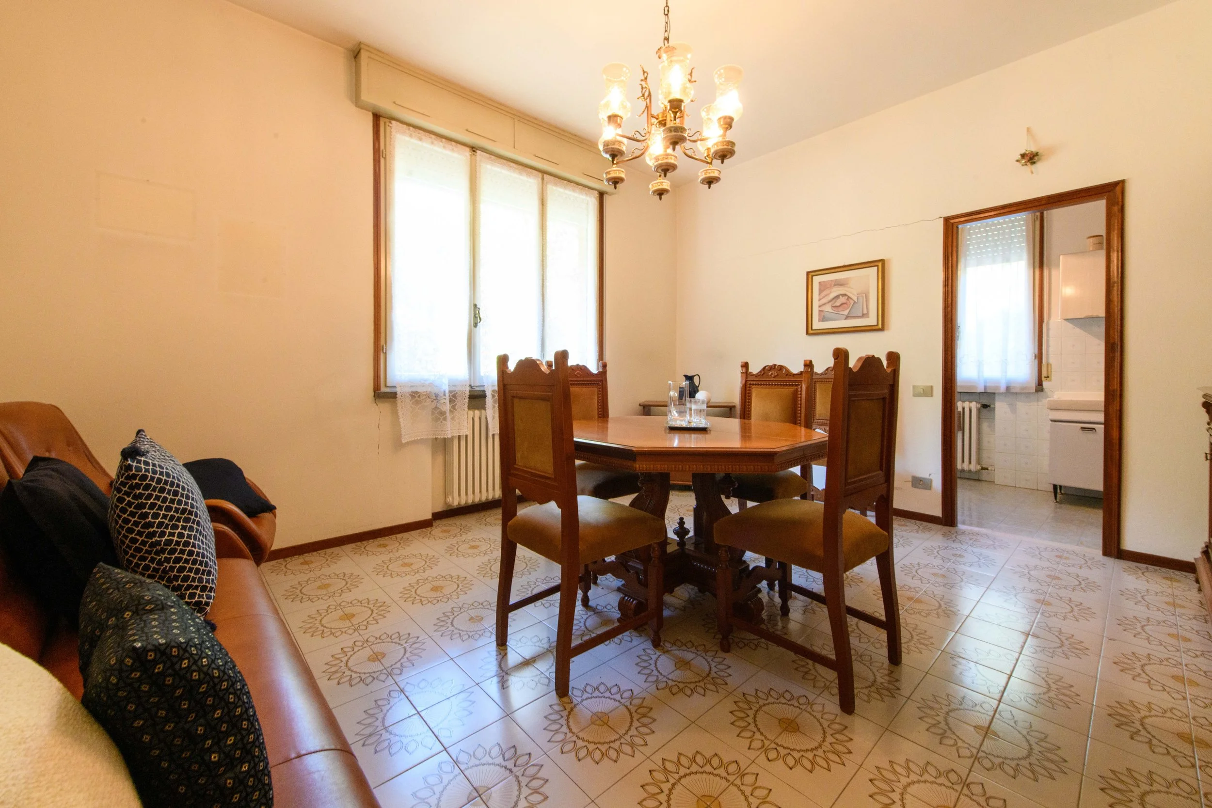 Sala da pranzo con tavolo di legno, sedie imbottite, lampadario classico e divano marrone con cuscini decorativi. Finestra con tende di pizzo, pavimento piastrellato con motivi floreali e porta che conduce alla cucina.