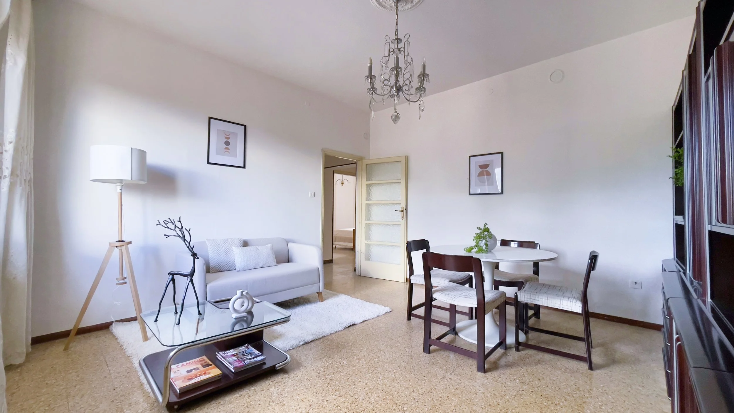 Soggiorno luminoso con divano bianco, lampada moderna, tavolino con decorazioni, tavolo da pranzo e sedie, quadri alle pareti e pavimento in legno chiaro. Arredamento elegante e minimalista.