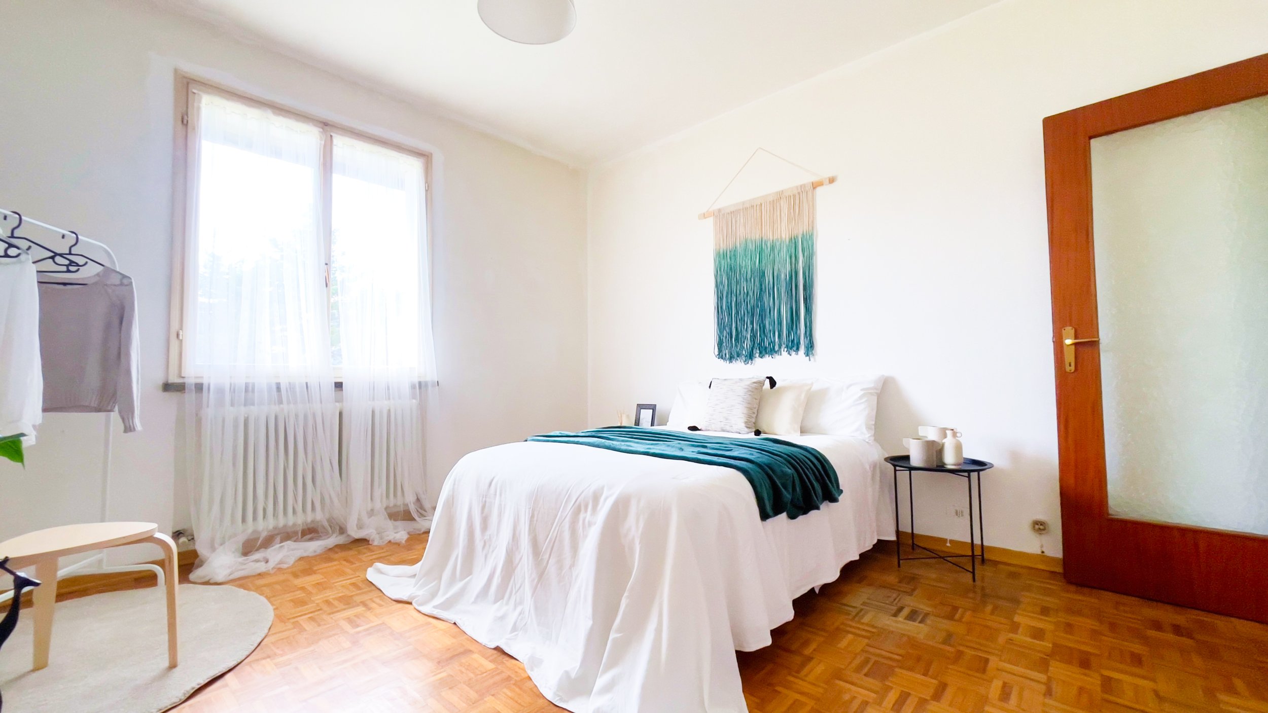 Camera da letto luminosa con letto matrimoniale, coperta verde e decorazione a parete in tessuto. Tavolino nero a lato del letto, vestiti appesi e grande finestra con tende bianche.