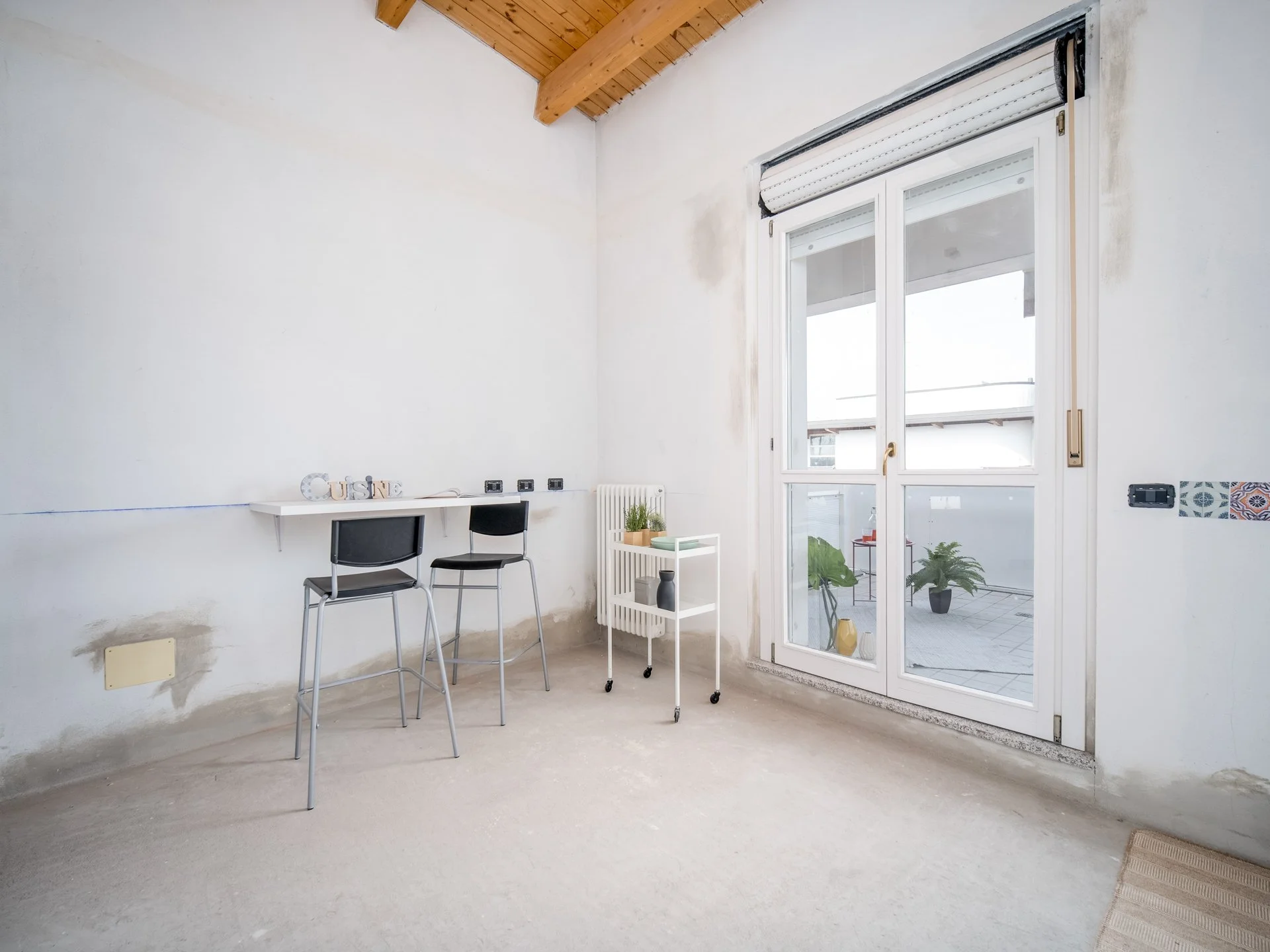 Interno di una stanza luminosa con pareti bianche e soffitto in legno. Due sgabelli sono posizionati davanti a un piccolo tavolo a muro. Vicino c'è un mobiletto bianco con piante sopra. Una grande finestra si apre su un patio esterno con piante in va