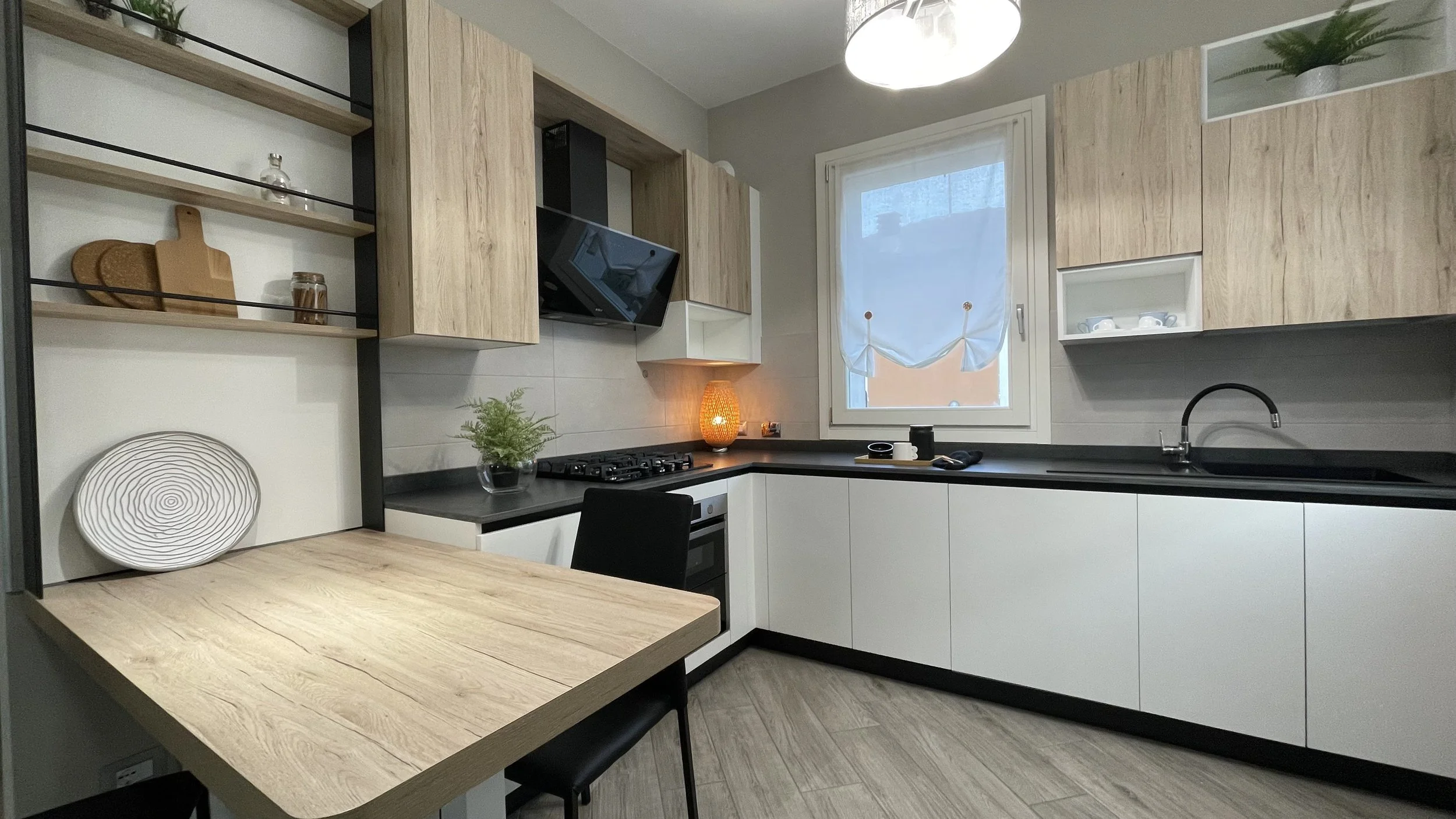 Cucina moderna con mobili in legno chiaro, piano di lavoro nero, fornelli a gas, cappa, lavandino nero, tavolo a penisola, mensole con decorazioni e una finestra con tende bianche.