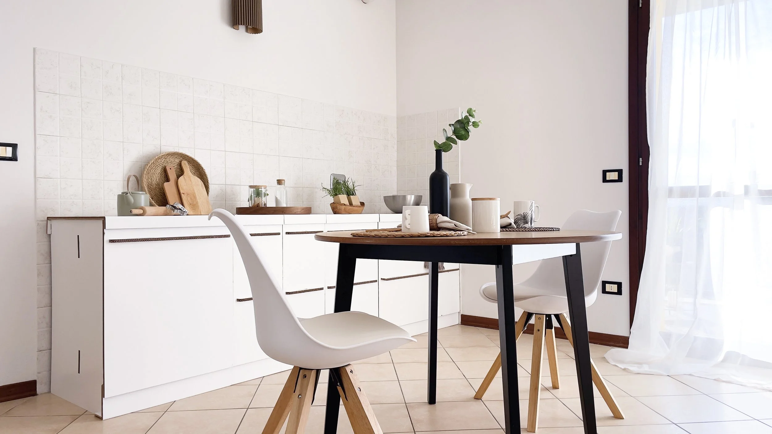 Cucina moderna con tavolo rotondo in legno, sedie bianche con gambe in legno, cassetti bianchi, decorazioni e piante, luce naturale da una finestra a destra.