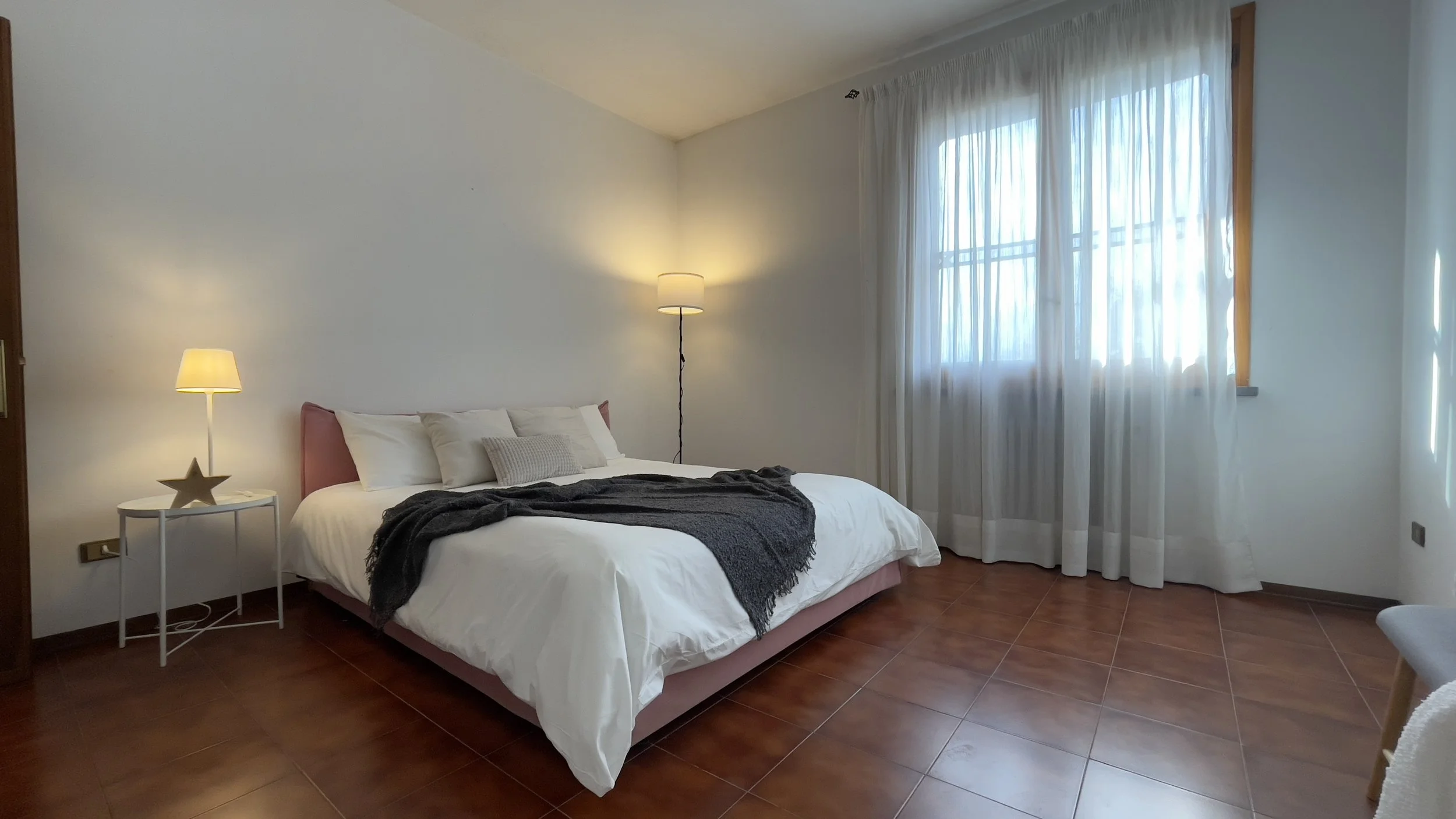 Camera da letto moderna con letto matrimoniale, coperta scura, comodino con lampada e finestra con tende leggere.