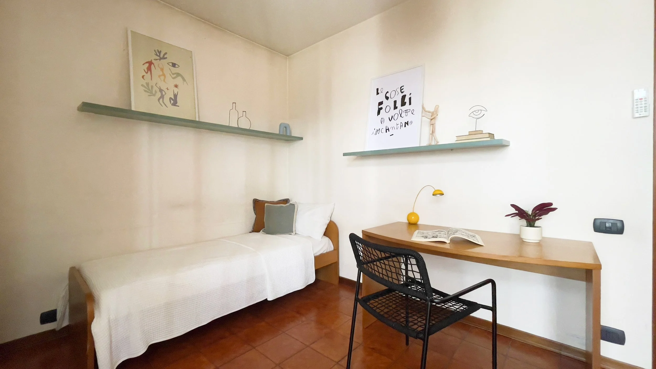 Camera minimalista con letto singolo e scrivania, decorata con opere d'arte e scaffali. C'è una sedia nera, una lampada gialla e una pianta in un vaso bianco. Le pareti sono di colore neutro e il pavimento è in piastrelle marroni.