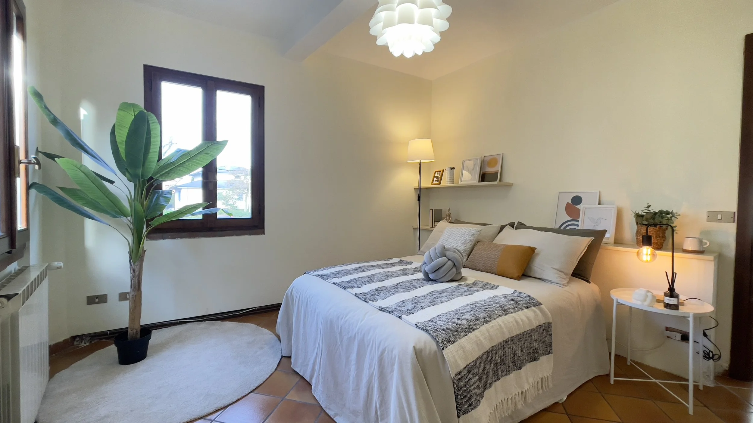 Camera da letto luminosa con letto matrimoniale, lampada, pianta, finestre e arredi moderni.