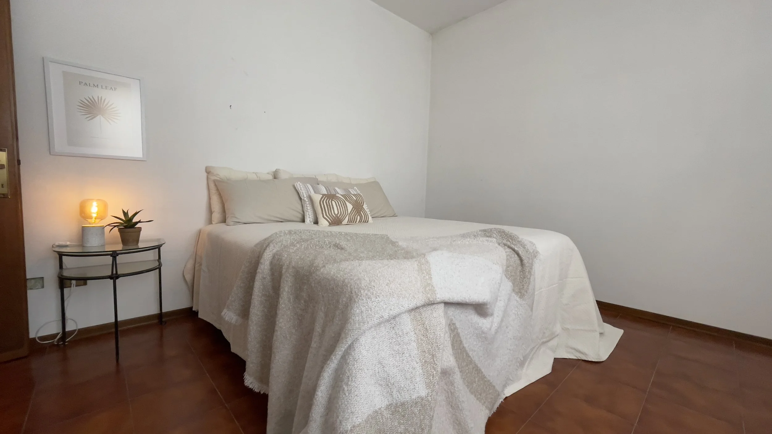 Camera da letto minimalista con letto matrimoniale, cuscini beige, coperta, tavolino con lampada accesa e pianta, quadro "Palm Leaf" sulla parete bianca.