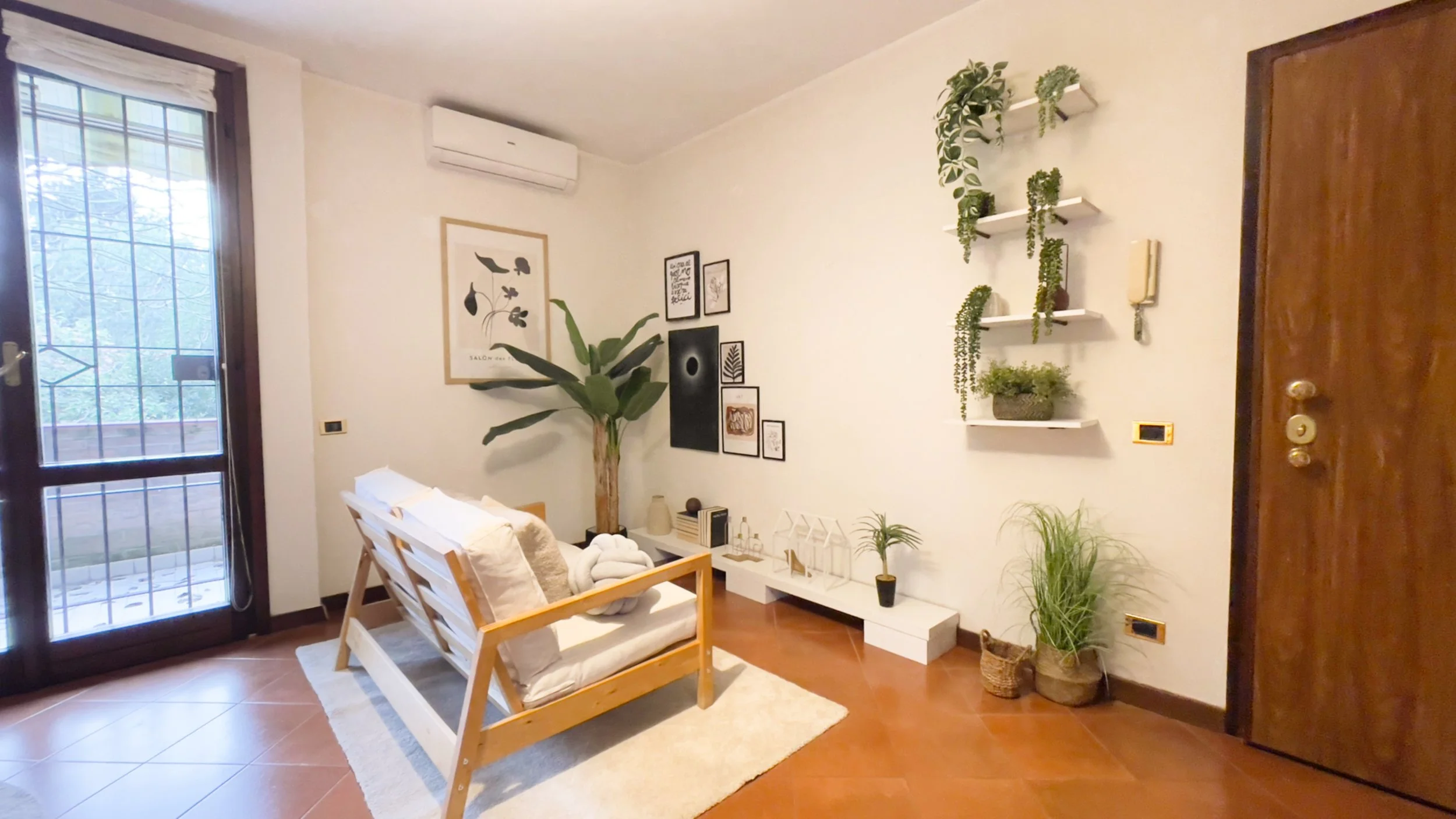 Soggiorno con arredo minimalista con divano in legno e cuscini bianchi, piante verdi e quadri alle pareti. Tavolino bianco con decorazioni e luce naturale da una grande finestra.