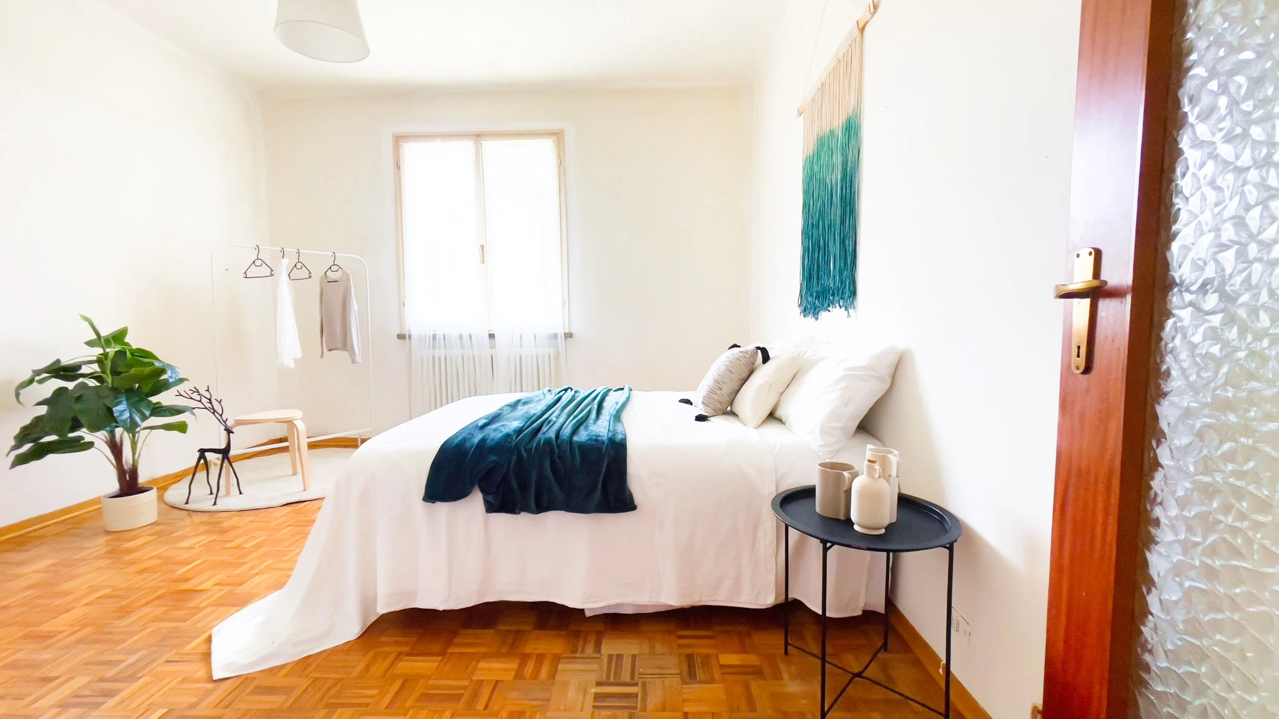 Camera da letto minimalista, con letto matrimoniale, coperta blu, comodino nero, tazze, pianta in vaso, appendiabiti con vestiti leggeri, sopra pavimento in parquet e decorazioni a parete.