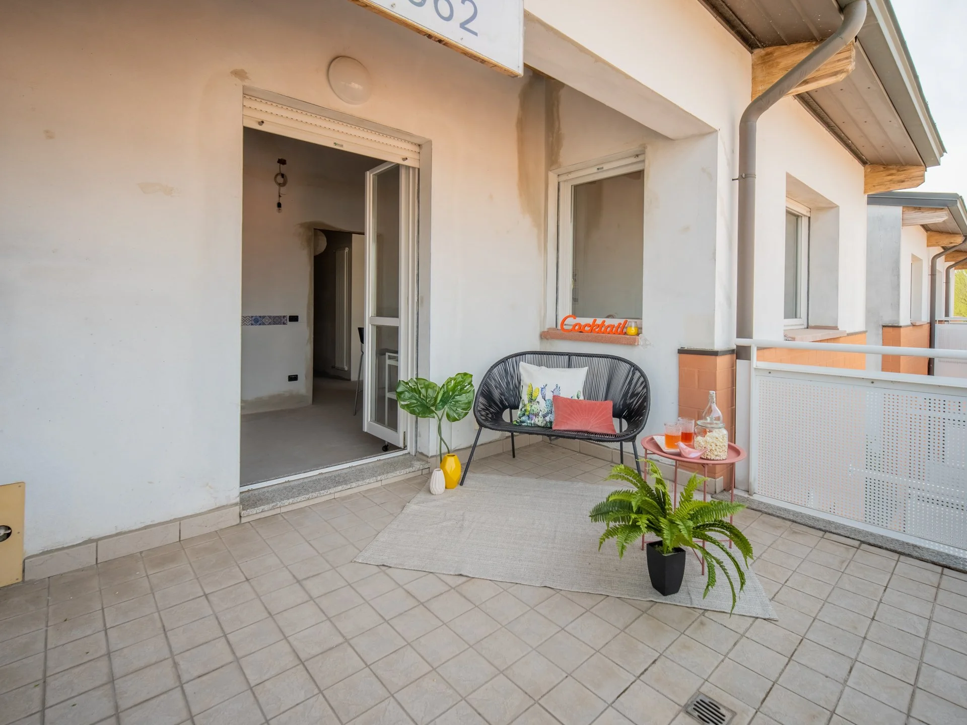 Terrazza con sedie, tavolino e pianta, bevande e snack, accanto a una porta che conduce all'interno.