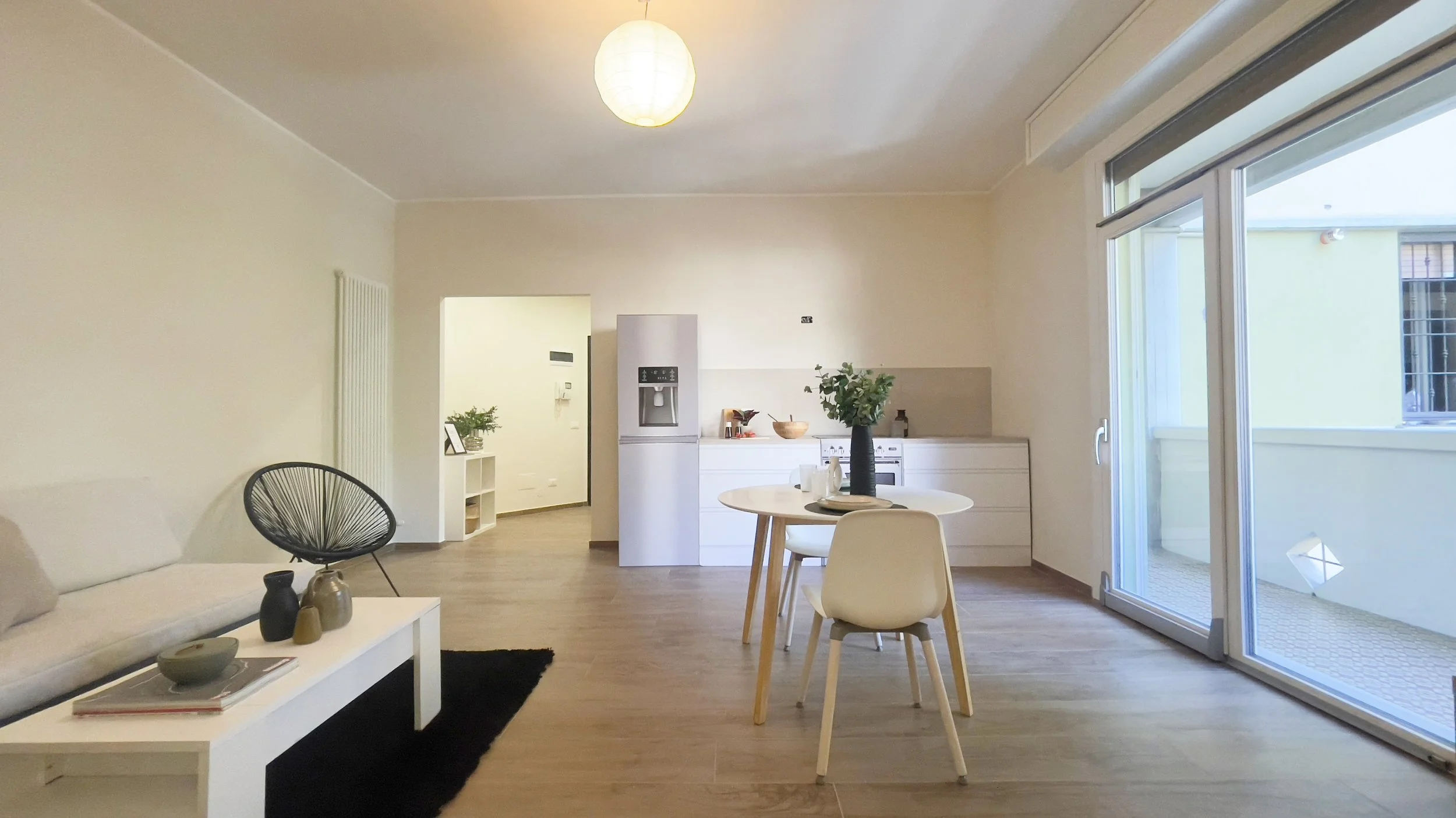 Soggiorno moderno con cucina a vista, tavolo da pranzo rotondo con sedie bianche, divano beige, tavolino bianco con decorazioni e grandi finestre con vista esterna.