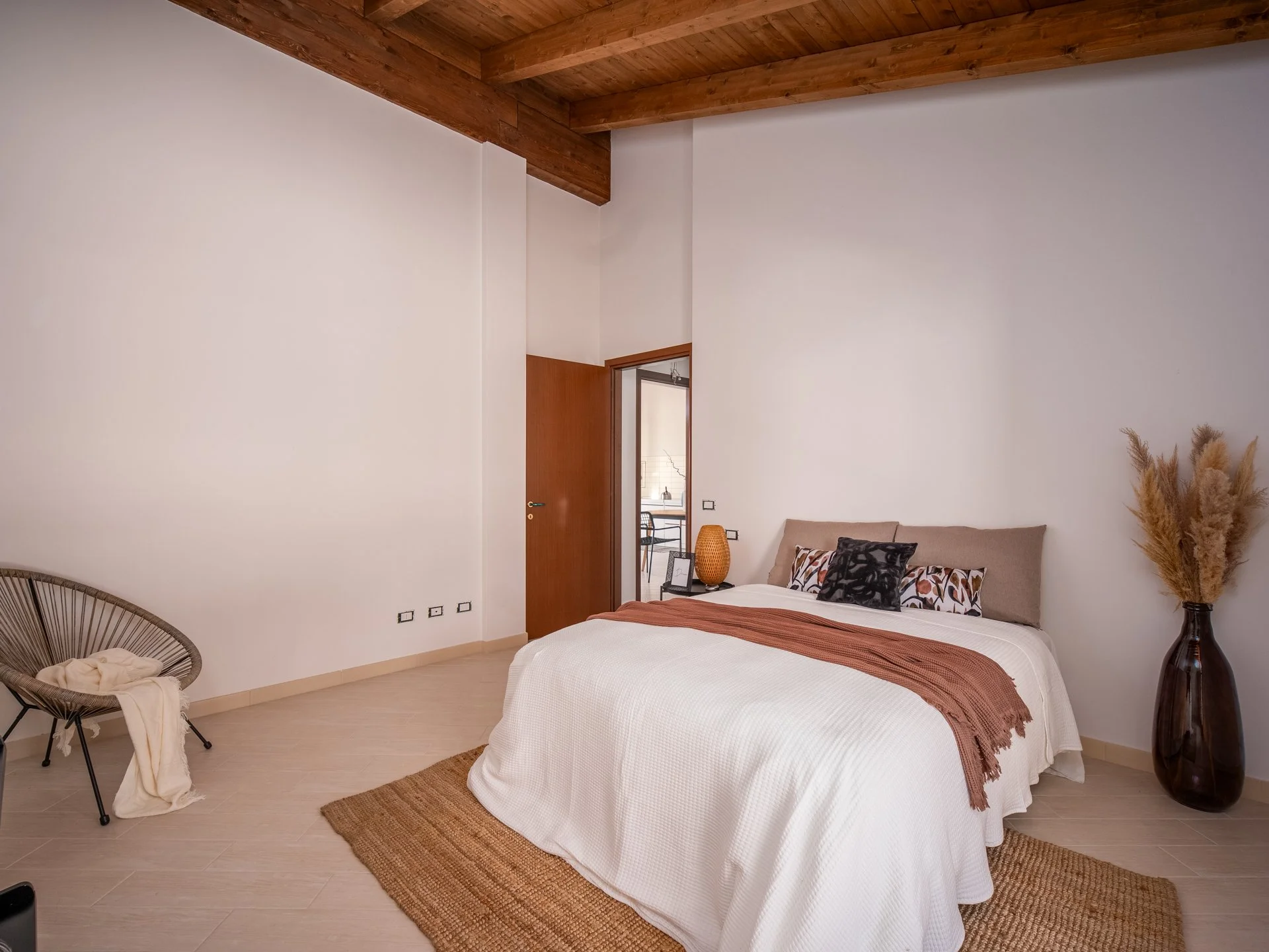 Camera da letto con letto matrimoniale, cuscini decorativi, coperta marrone, poltrona con coperta chiara, vasi decorativi e soffitto con travi in legno."