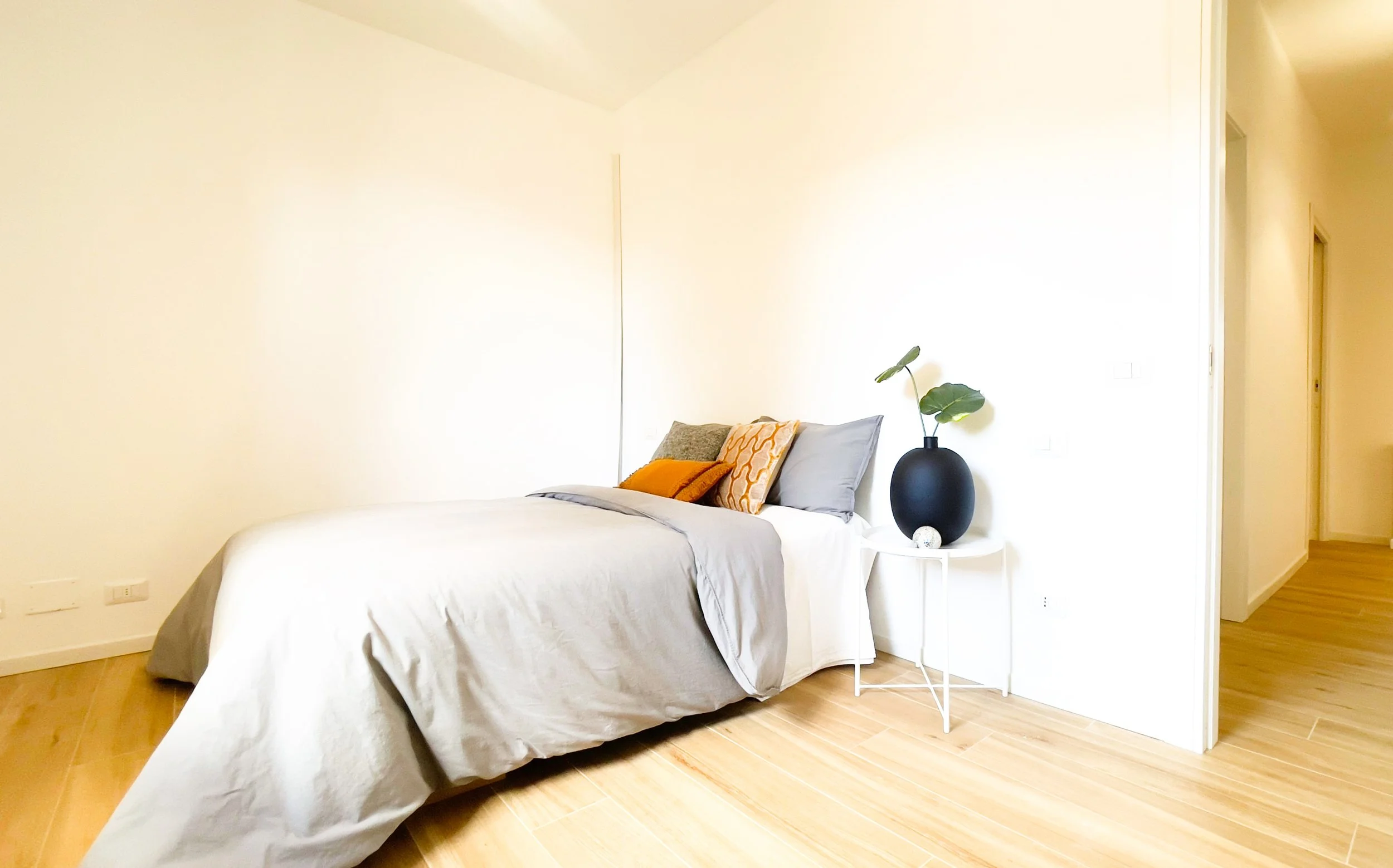 Camera da letto minimalista con letto matrimoniale, copriletto grigio, cuscini decorativi, tavolino bianco con vaso nero e pianta, pavimento in legno chiaro.