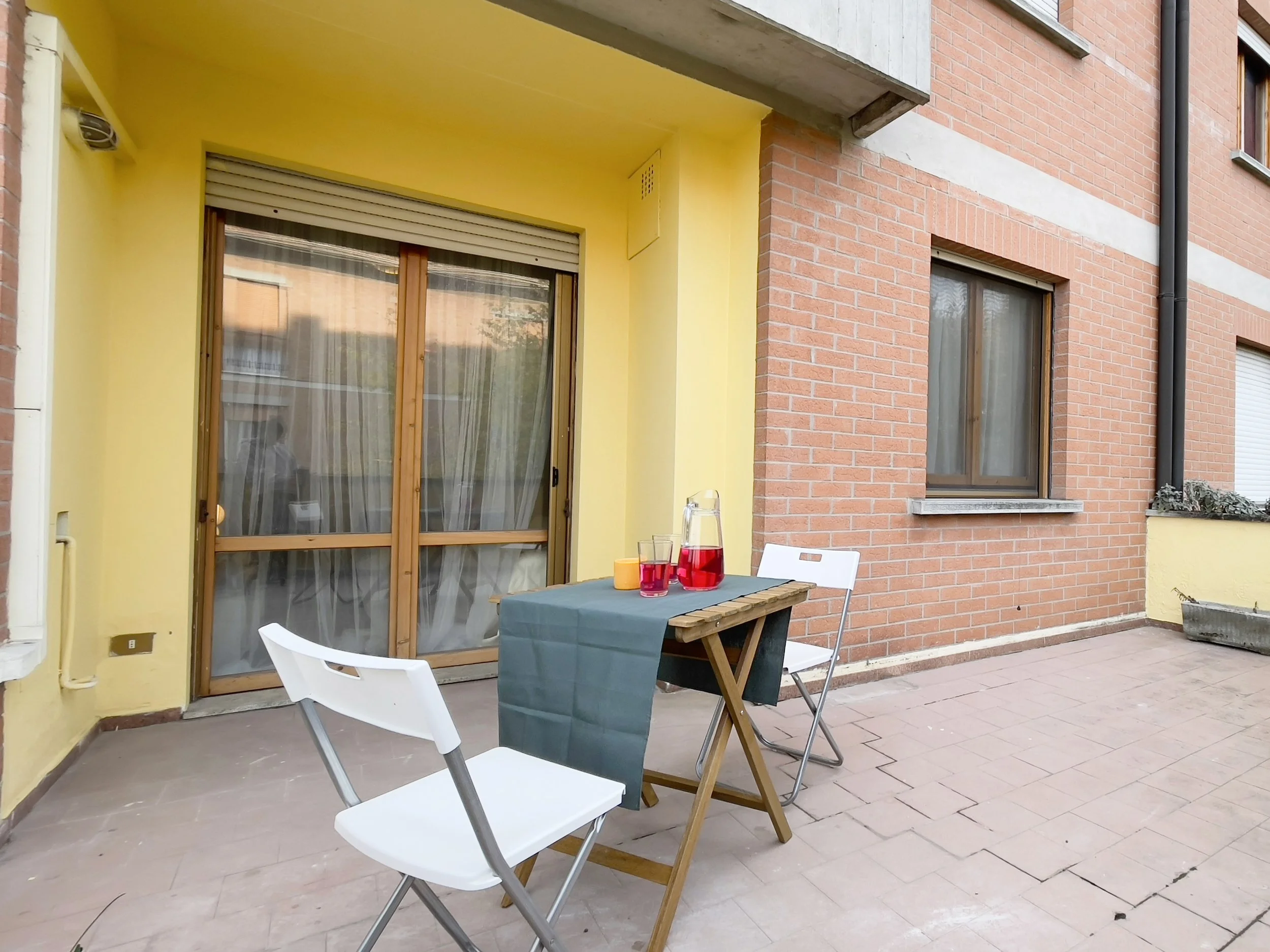 Terrazza con tavolino e sedie bianche, tovaglia blu, brocca di bevanda rossa e bicchieri, vicino a porta finestra su parete gialla e facciata in mattoni rossi.