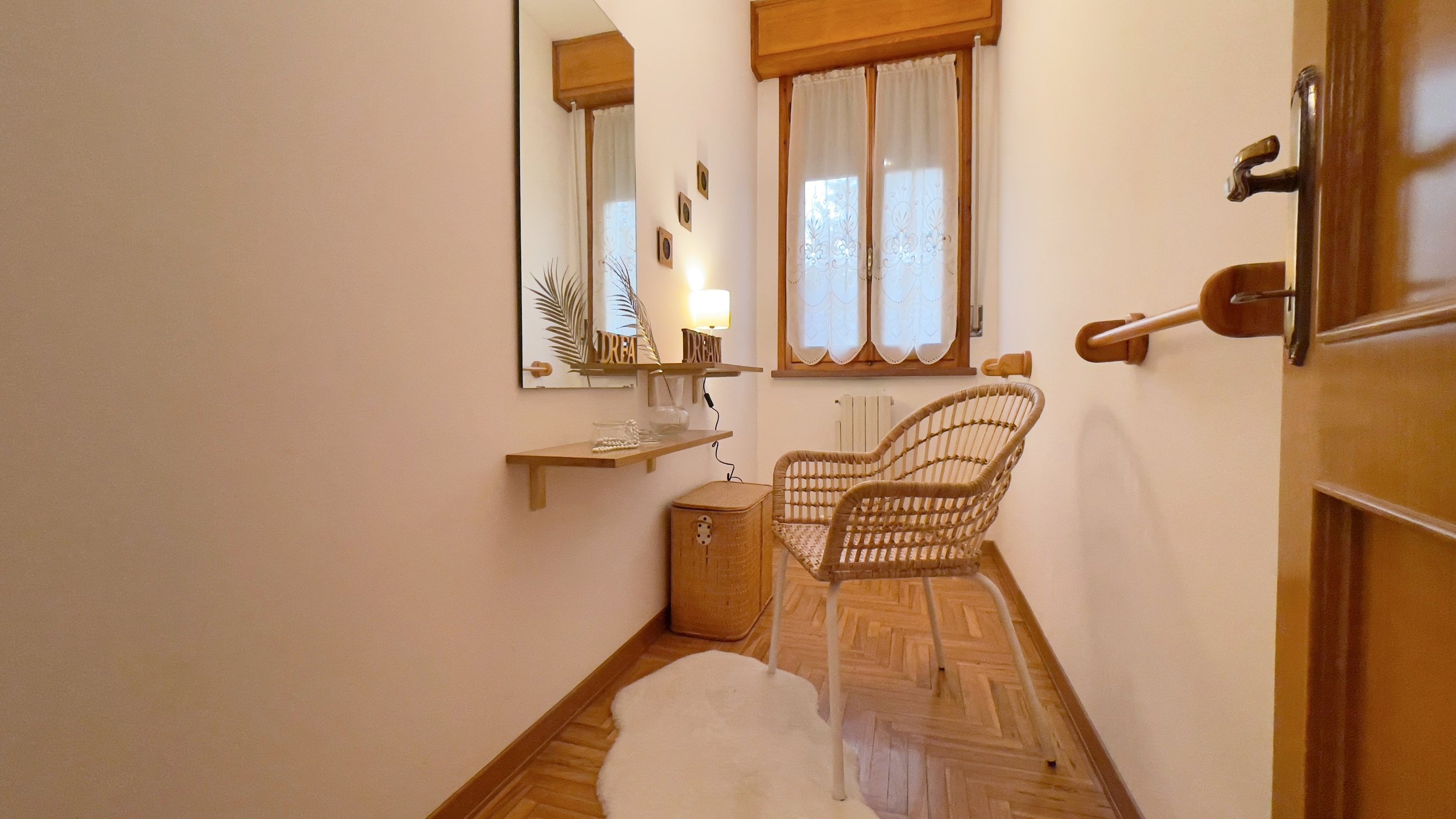 Piccola stanza con sedia di vimini, specchio a muro, mensola con decorazioni, finestra con tende bianche, pavimento in parquet e tappeto bianco.