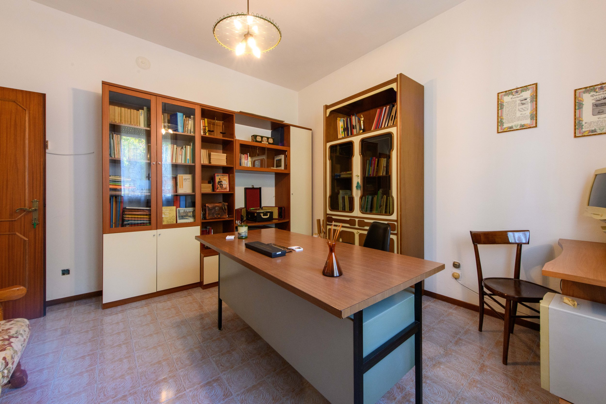 Ufficio con scrivania e libreria, sedia e computer, illuminato da una lampada a soffitto. Pareti bianche e pavimento a piastrelle.