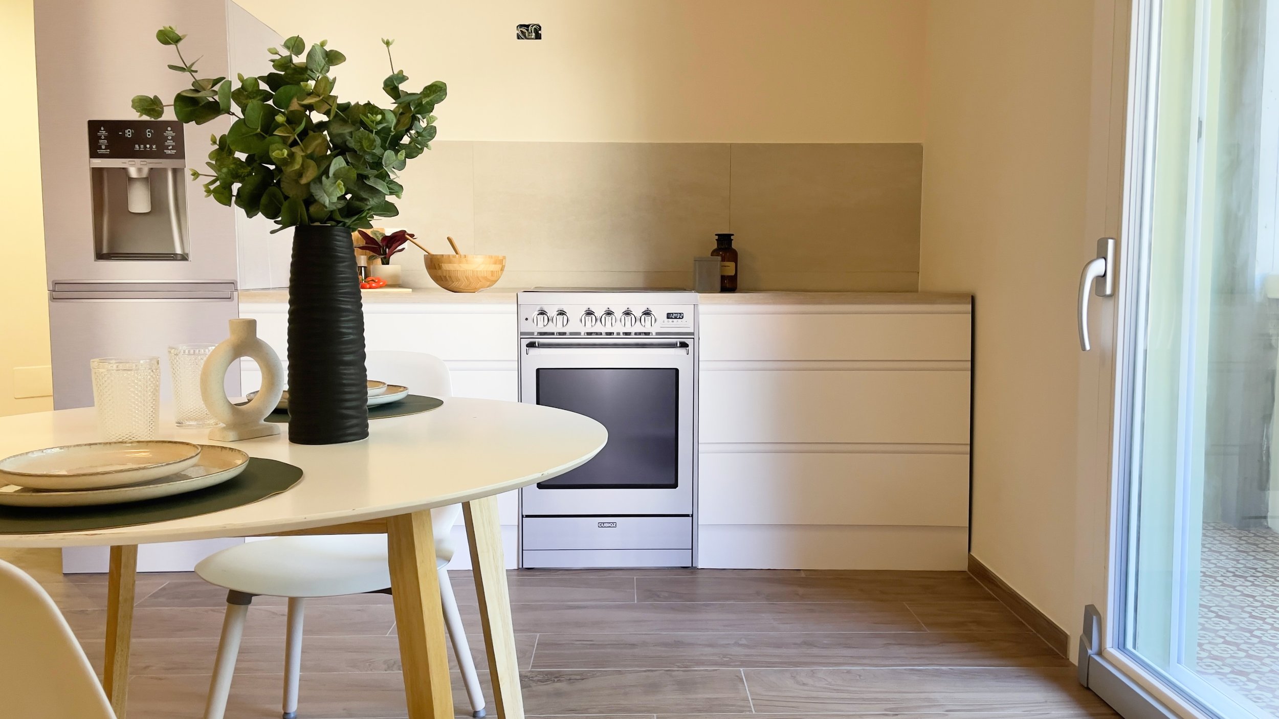 Cucina moderna con tavolo rotondo e sedie bianche, vaso con piante verdi, piatti e bicchieri, forno e frigorifero, pavimento in legno, e porta vetro scorrevole.