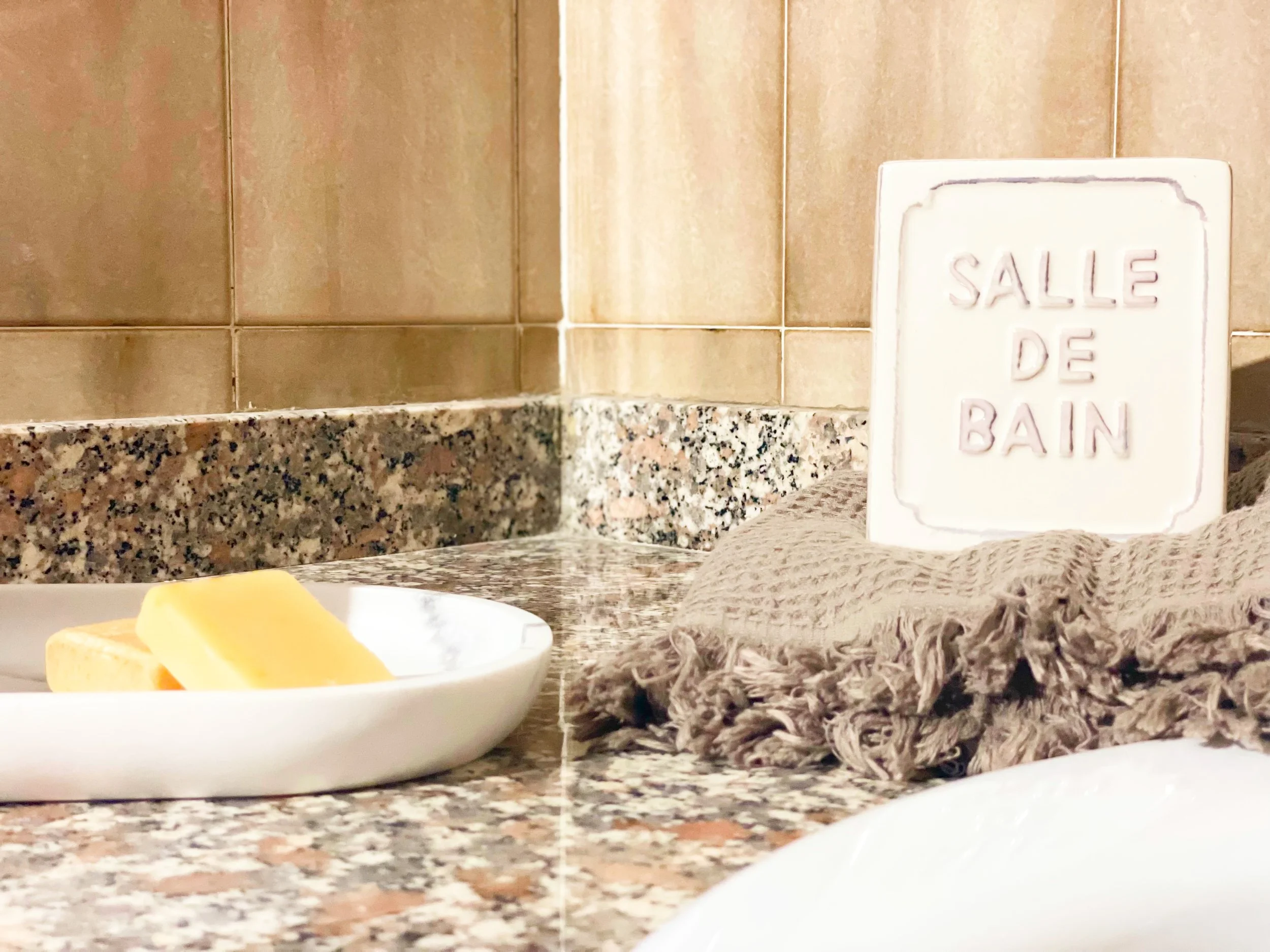 Piano del bagno con sapone su piattino bianco e targa con scritta "SALLE DE BAIN" su asciugamano beige a frange.