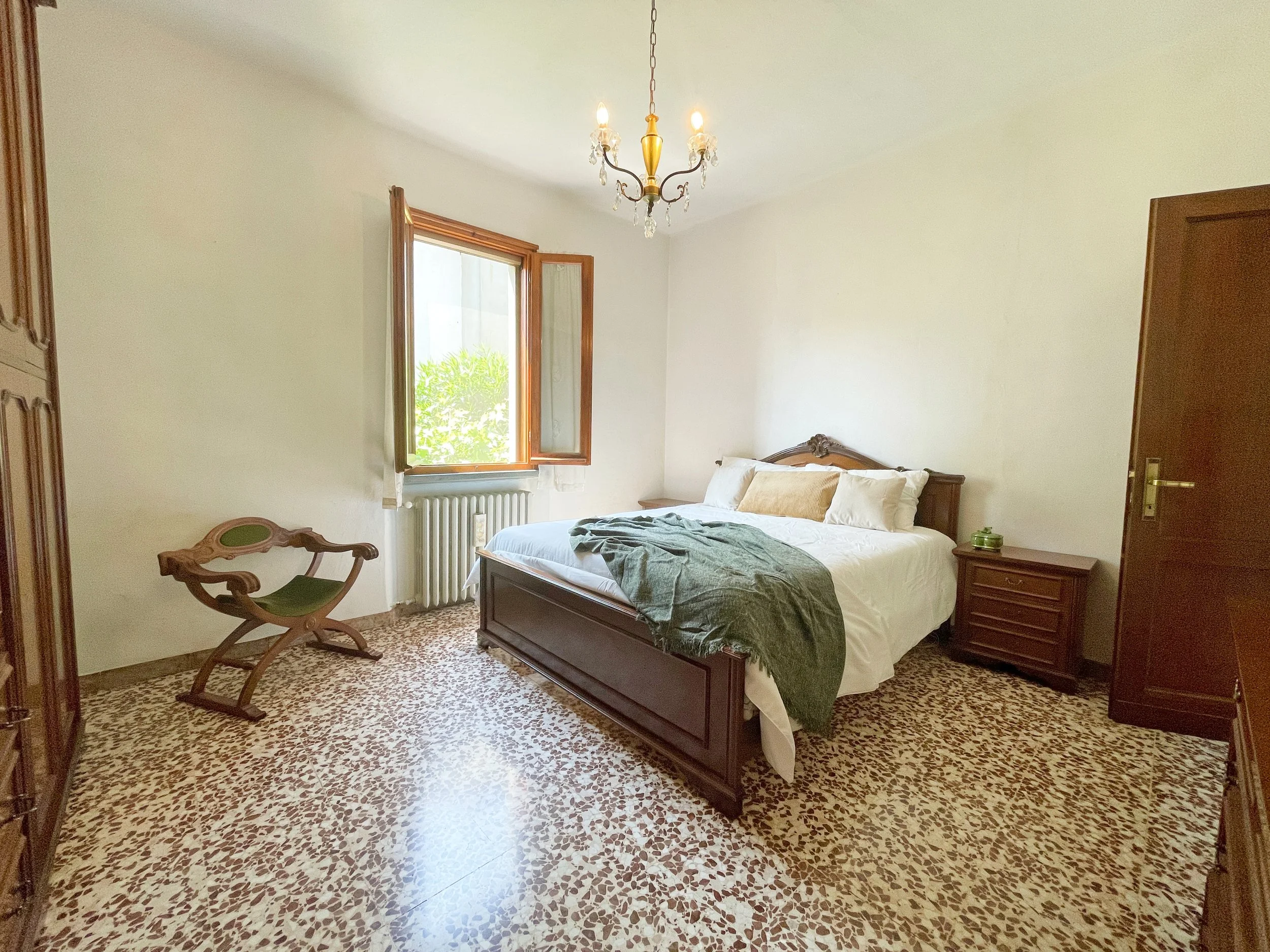 Camera da letto con letto matrimoniale, cassettiera, sedia in legno, finestra aperta e lampadario. Pavimento in piastrelle decorative, pareti bianche.