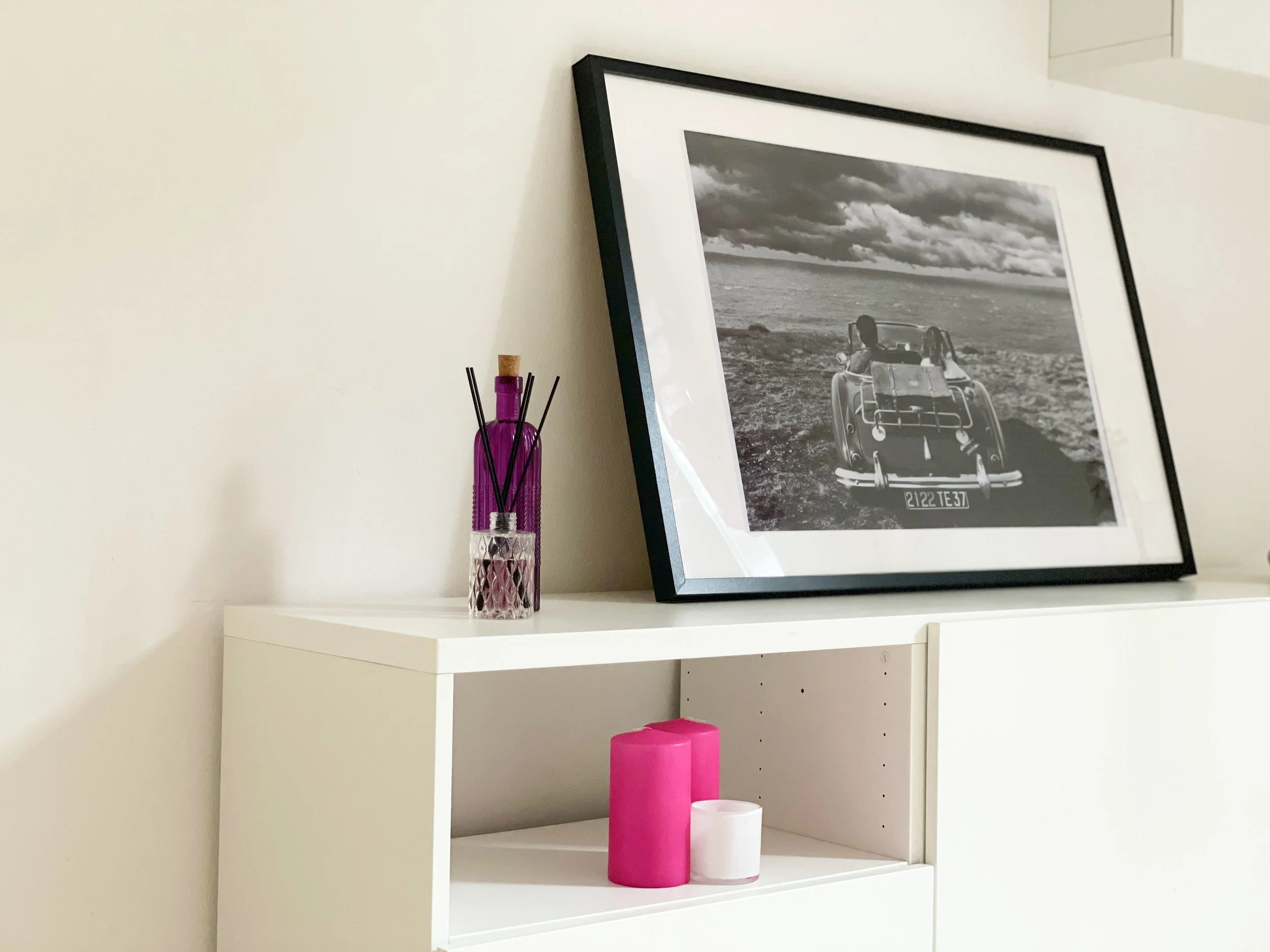 Interno di una stanza con una fotografia in bianco e nero di un'auto d'epoca in una cornice nera appoggiata su un mobile bianco. Accanto ci sono candele color rosa e bianco e un diffusore con bottiglia viola.