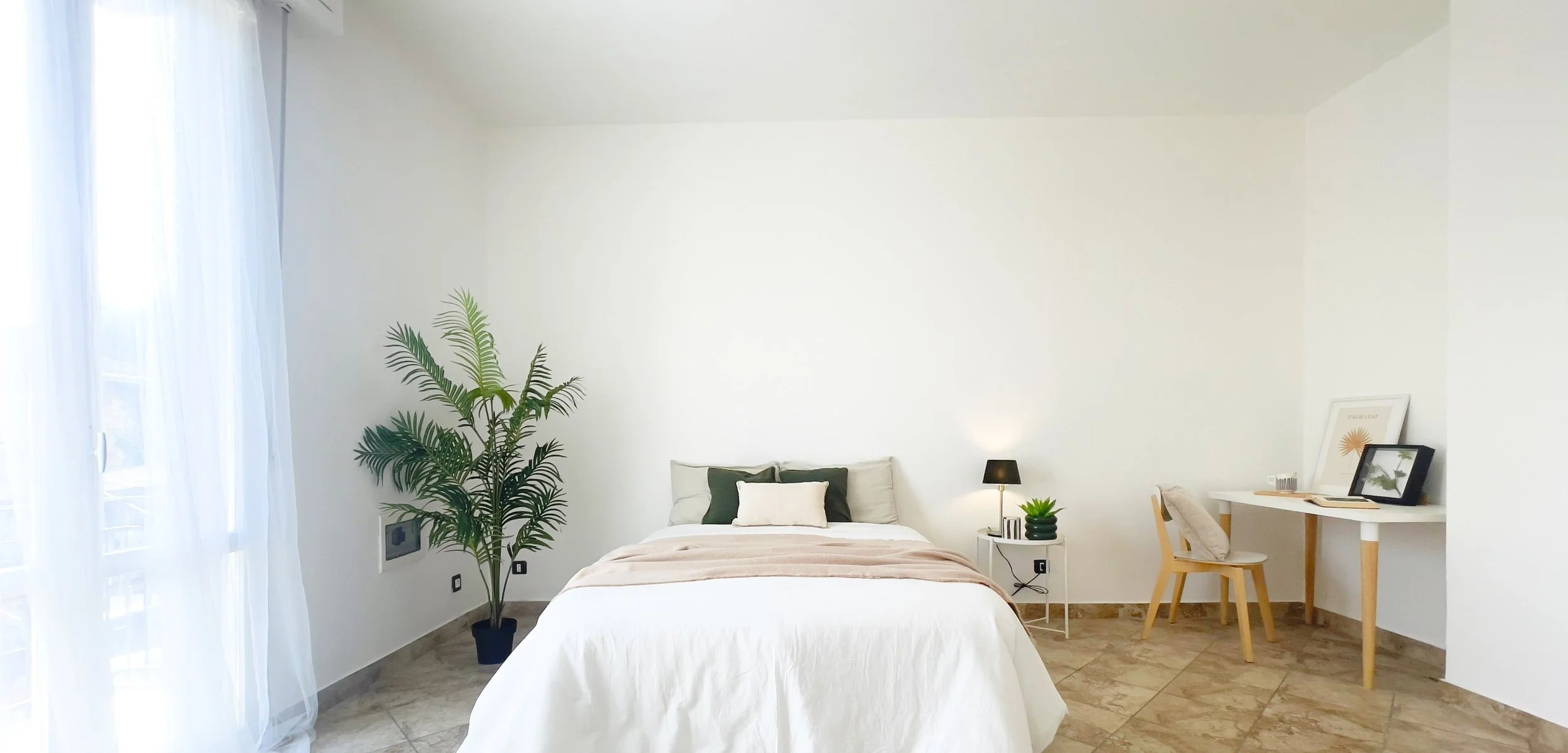 Camera da letto moderna con letto matrimoniale, pianta ornamentale, finestra con tende leggere, tavolino con lampada e pianta, scrivania con sedia, decorazione minimalista.