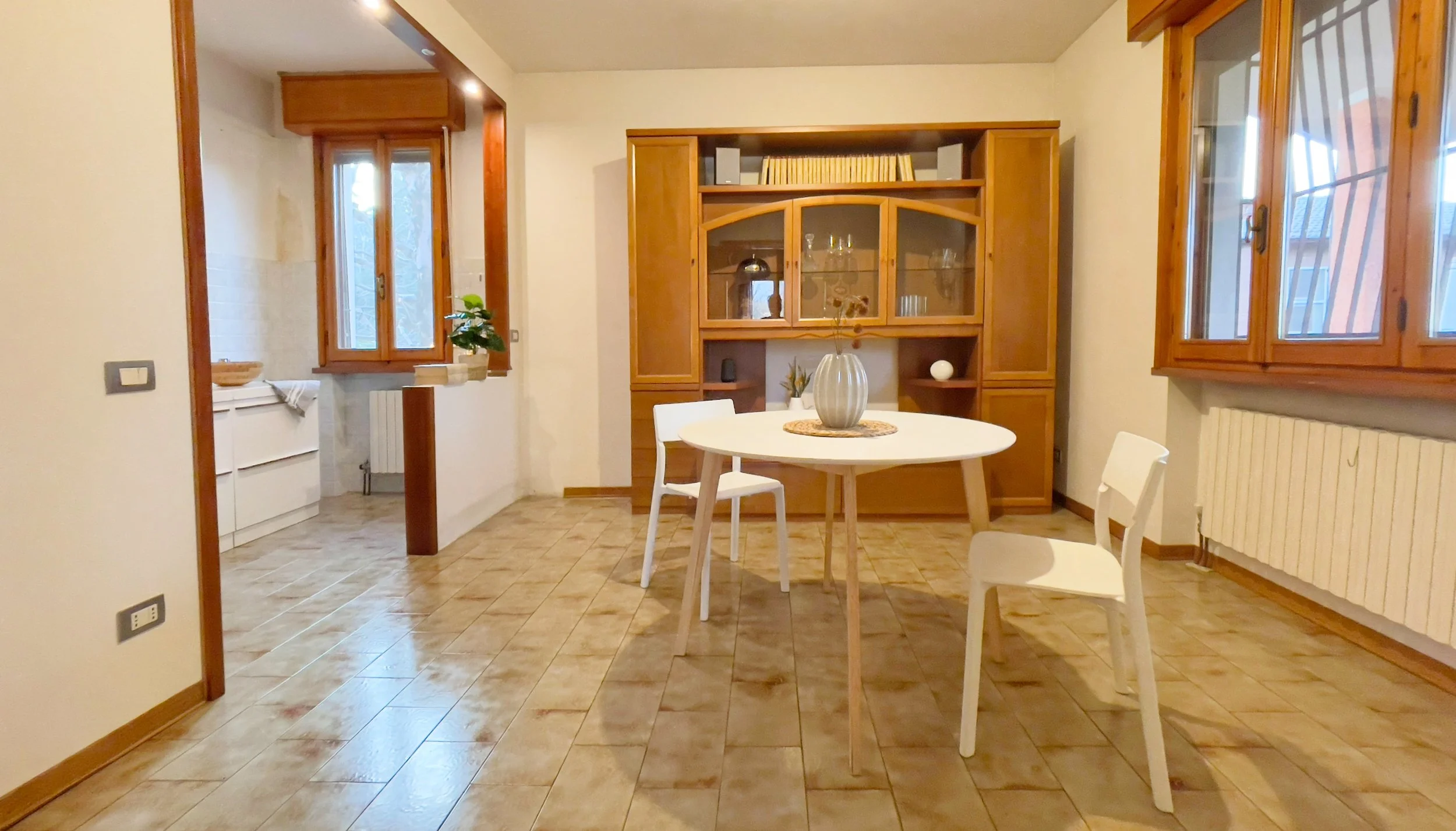 Interno di una sala da pranzo con tavolo rotondo e due sedie bianche, mobile in legno sullo sfondo, pavimento in piastrelle, grande finestra e radiatore sotto la finestra.
