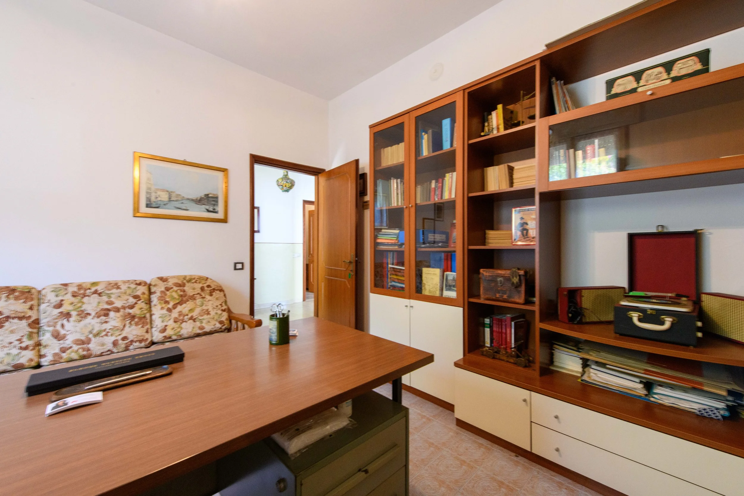 Studio con scrivania di legno, sedia con fodera a motivi floreali, libreria piena di libri e oggetti decorativi, porta aperta che conduce in un'altra stanza e quadro appeso alla parete.