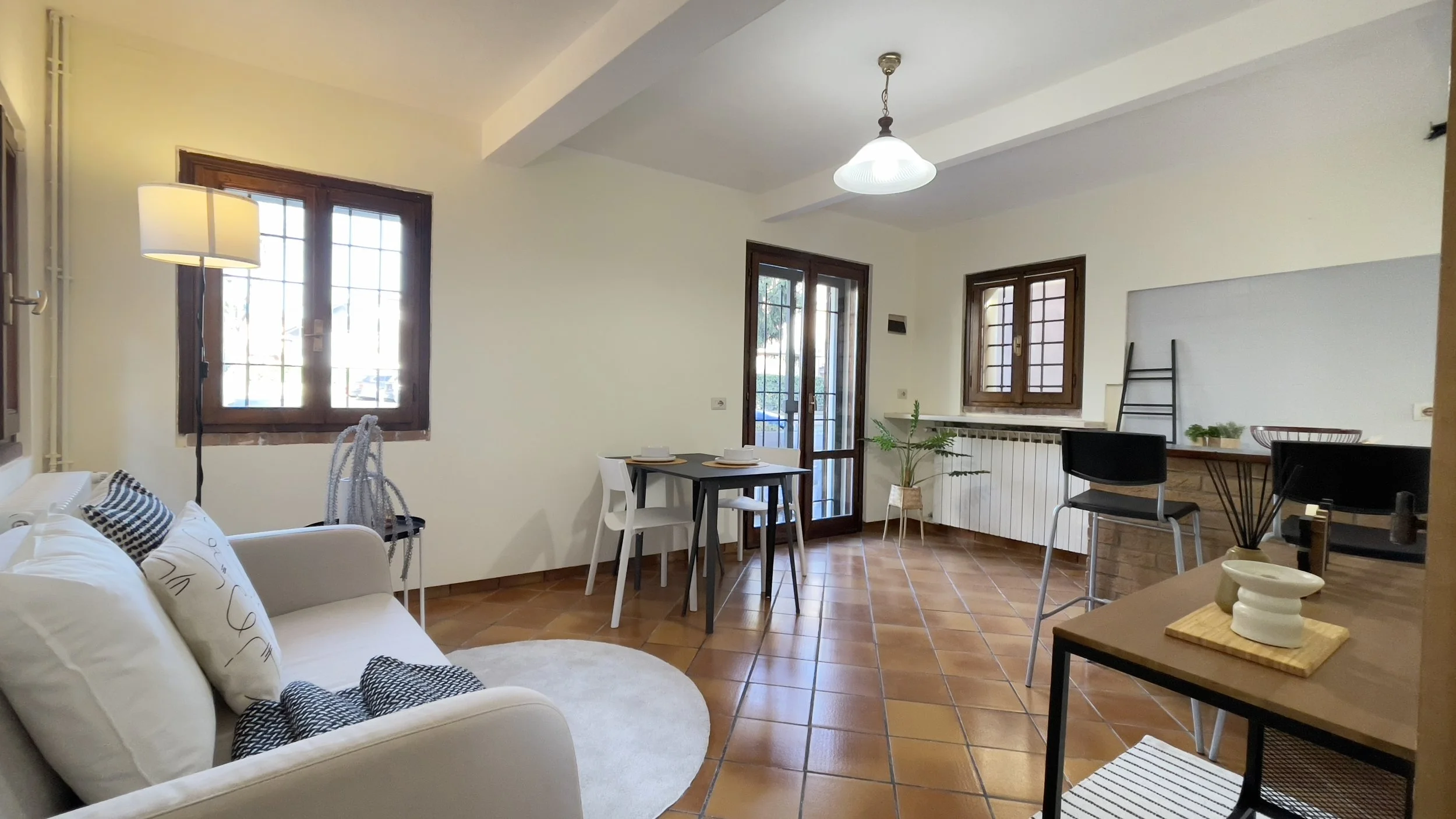 Soggiorno luminoso con pavimento in piastrelle marroni, divano con cuscini bianchi e neri, tavolo da pranzo nero con sedie bianche, e finestre con infissi in legno. Arredamento minimalista e moderno.