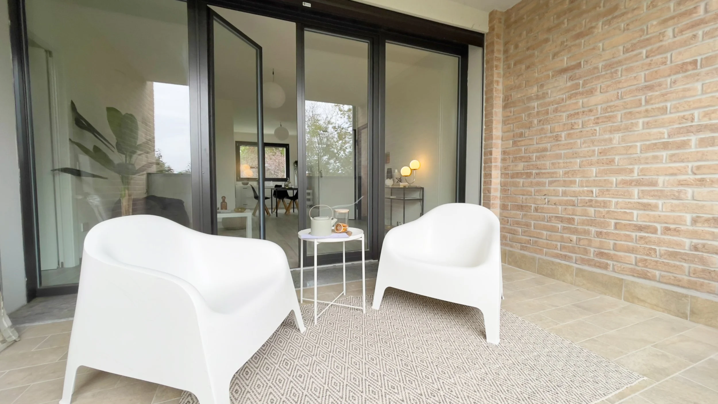 Due sedie bianche su un balcone con una parete di mattoni, tavolino bianco, e porte scorrevoli in vetro che conducono all'interno della casa.