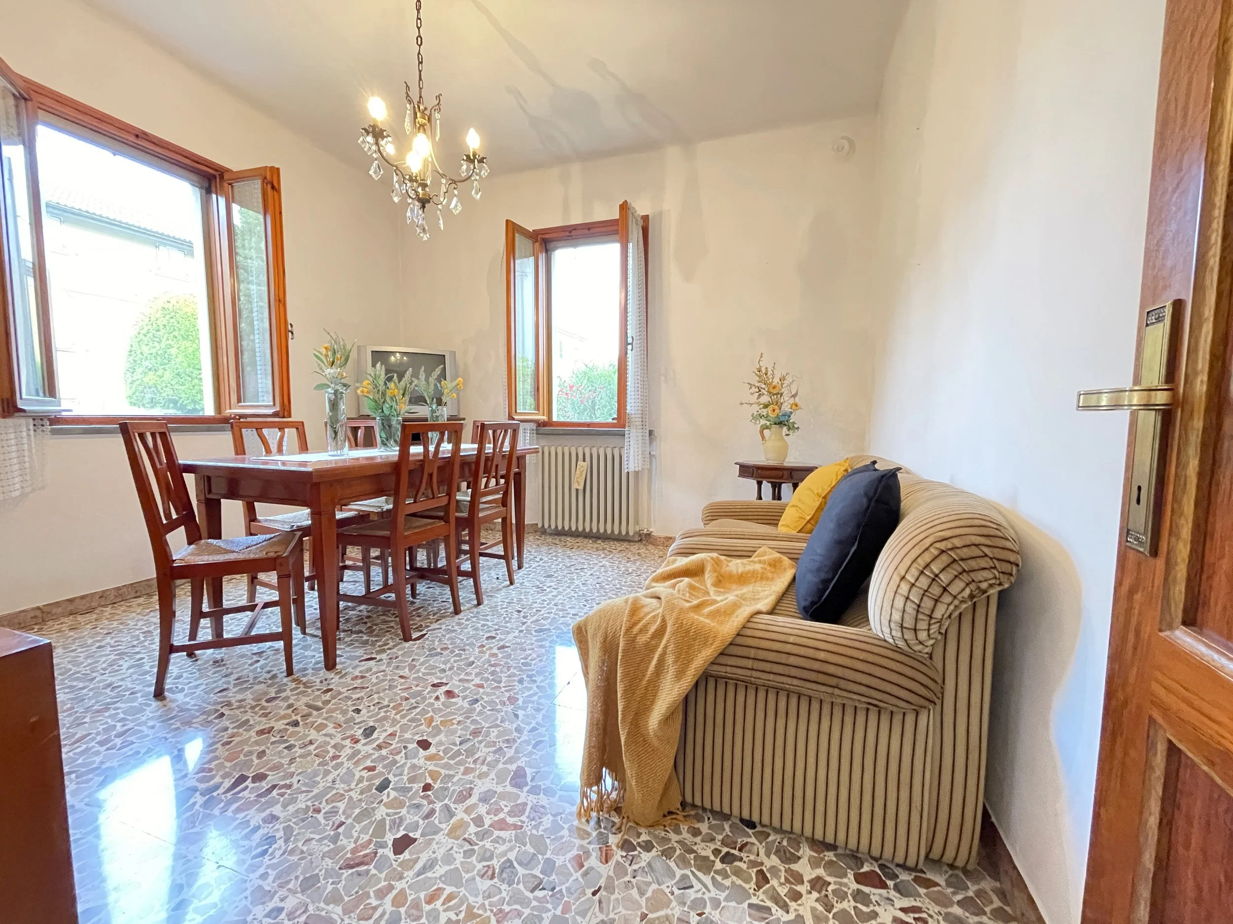 Sala da pranzo con tavolo di legno, sedie, divano a righe, cuscini colorati, vasi di fiori, grande finestra e lampadario.