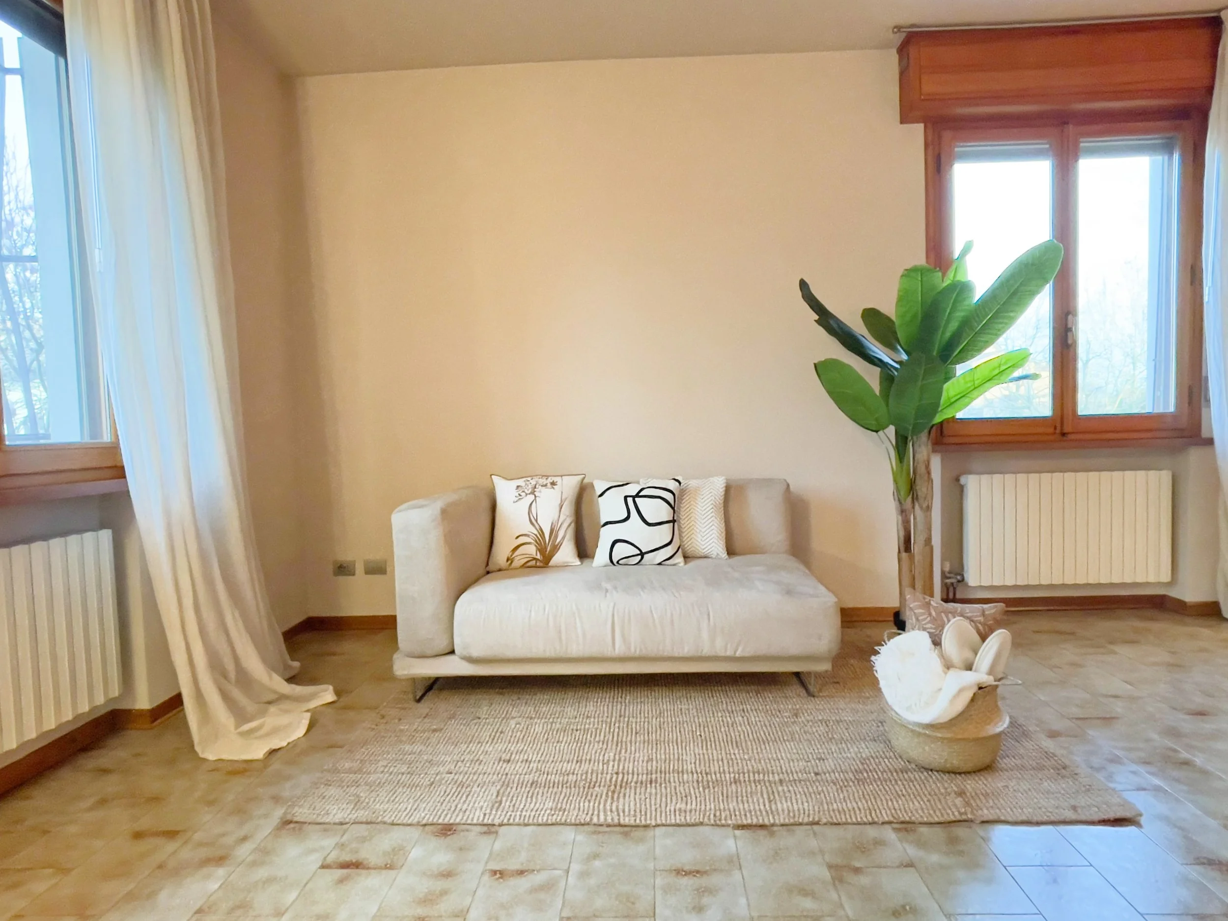 Soggiorno minimalista con divano beige, piante verdi, cuscini decorativi, tappeto intrecciato, e ampie finestre con tende bianche.