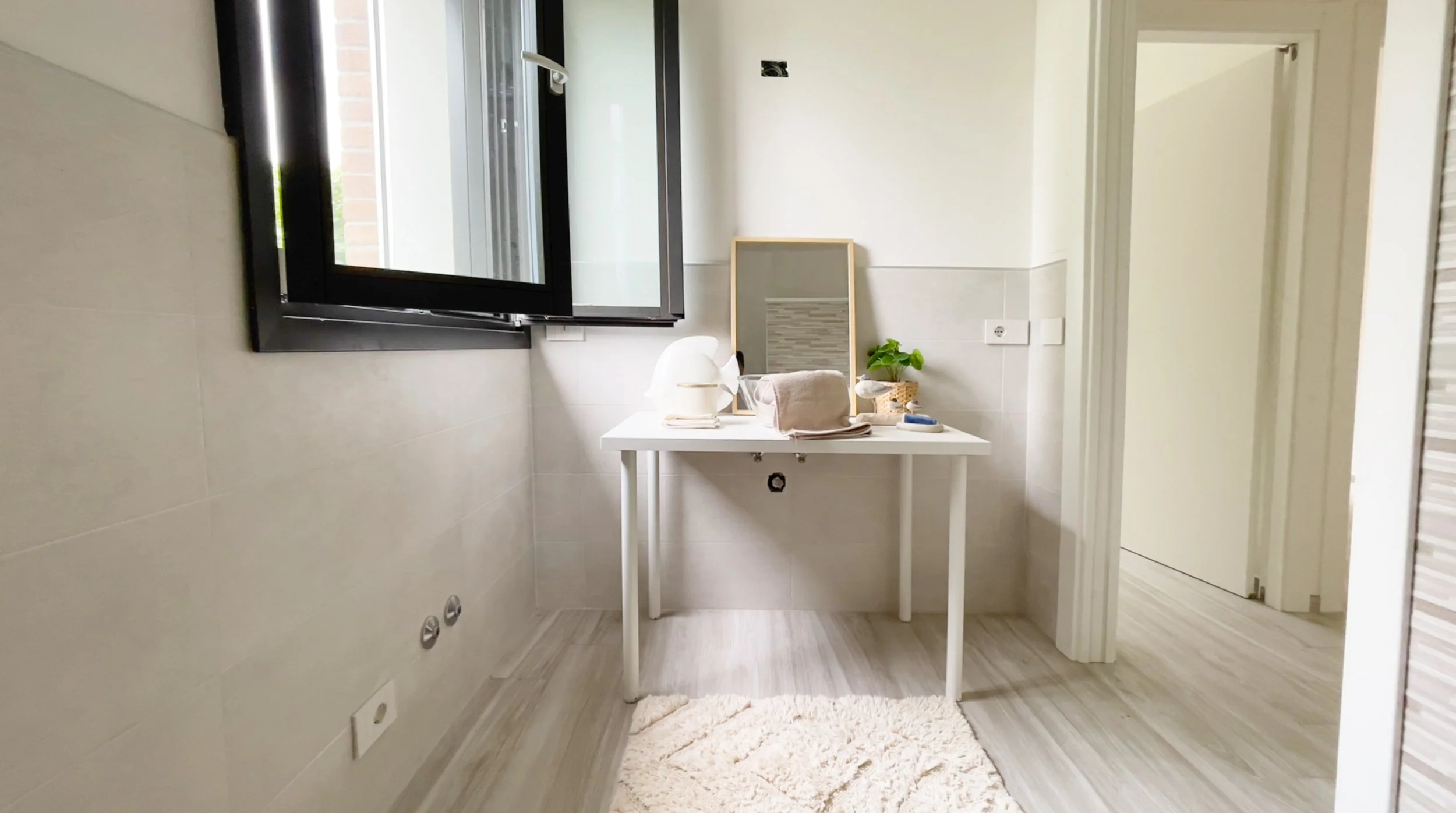 Interno di un bagno luminoso con finestra aperta, tavolo bianco con specchio, lampada, asciugamano e pianta. Pavimento in legno chiaro e tappeto morbido chiaro.