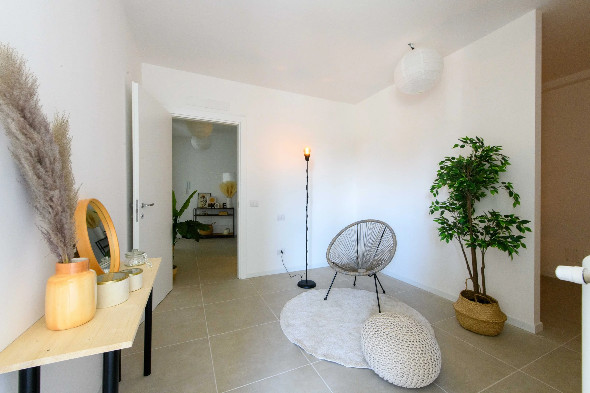 Stanza moderna con arredamento minimalista, comprende una sedia di design, un cuscino intrecciato su un tappeto chiaro, una pianta in vaso, una lampada da terra e un tavolino con uno specchio e decorazioni. Sullo sfondo si vede un'altra stanza.