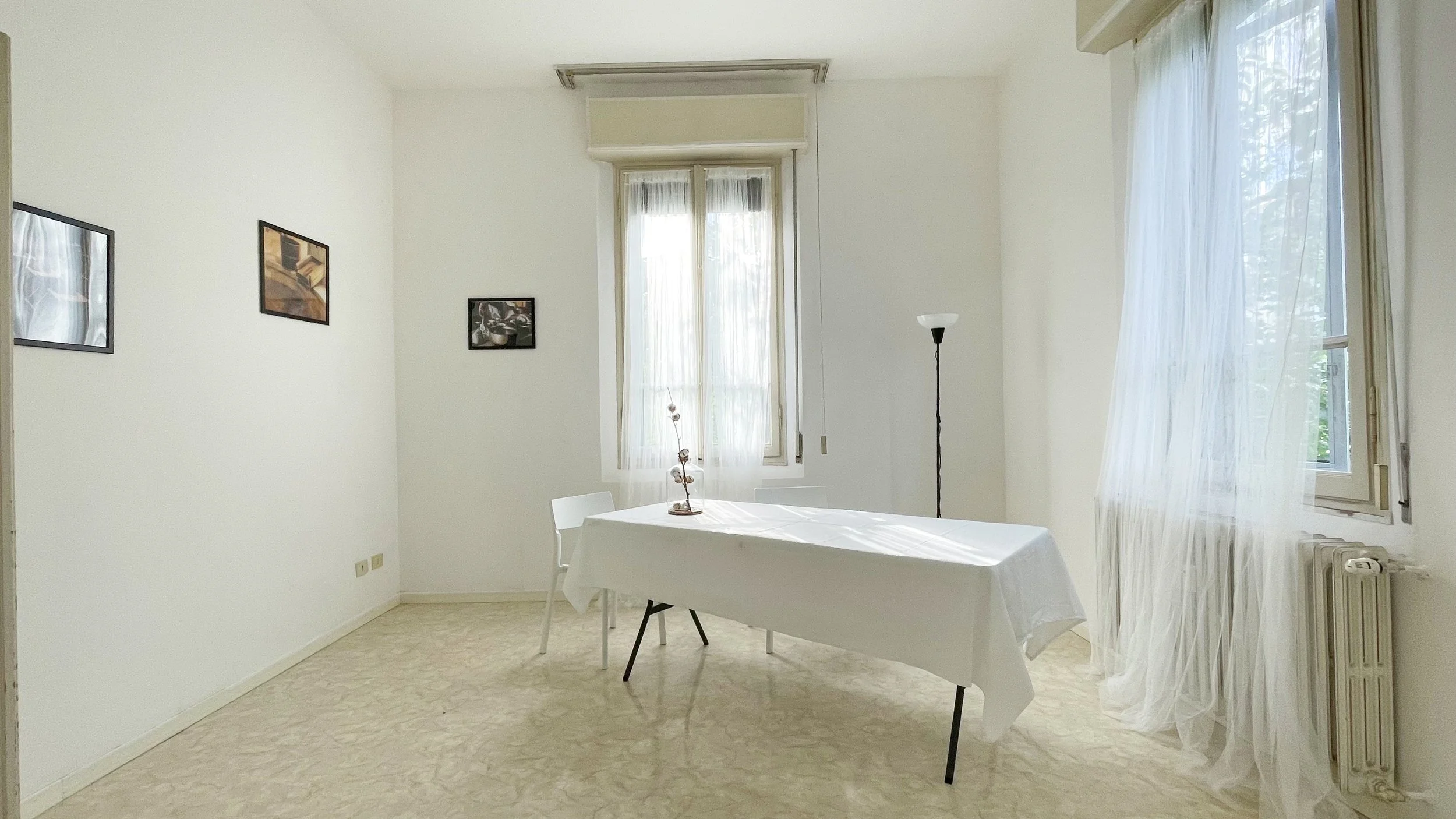 Interno di una stanza luminosa con tavolo rettangolare coperto da tovaglia bianca e sedie bianche. Ci sono quadri appesi alle pareti e una pianta ornamentale sopra il tavolo. Due finestre con tende leggere e un radiatore sono visibili. Una lampada a 