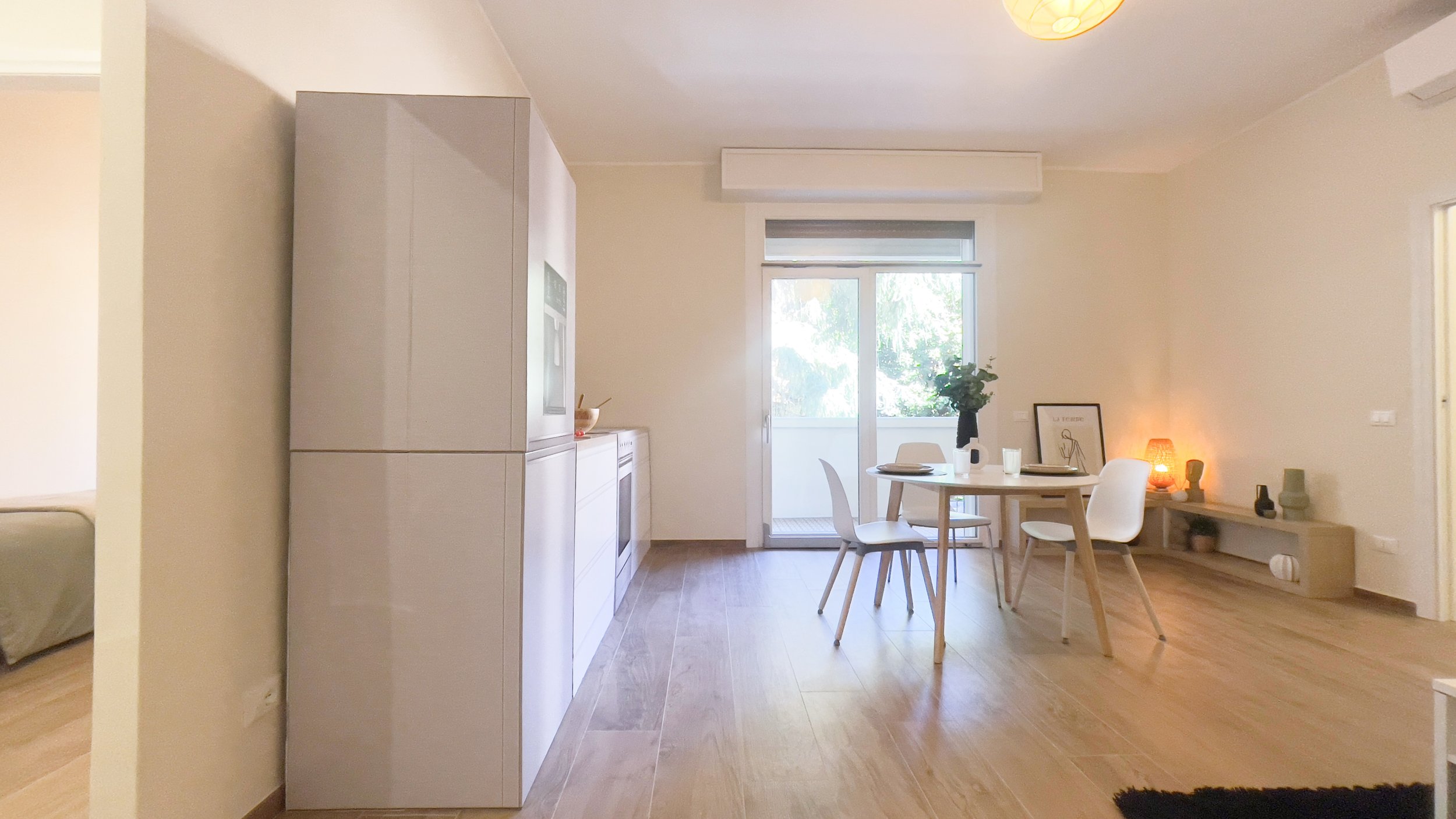 Sala da pranzo moderna con tavolo rotondo, sedie bianche, cucina a scomparsa e decorazioni minimaliste, illuminata da una grande finestra.