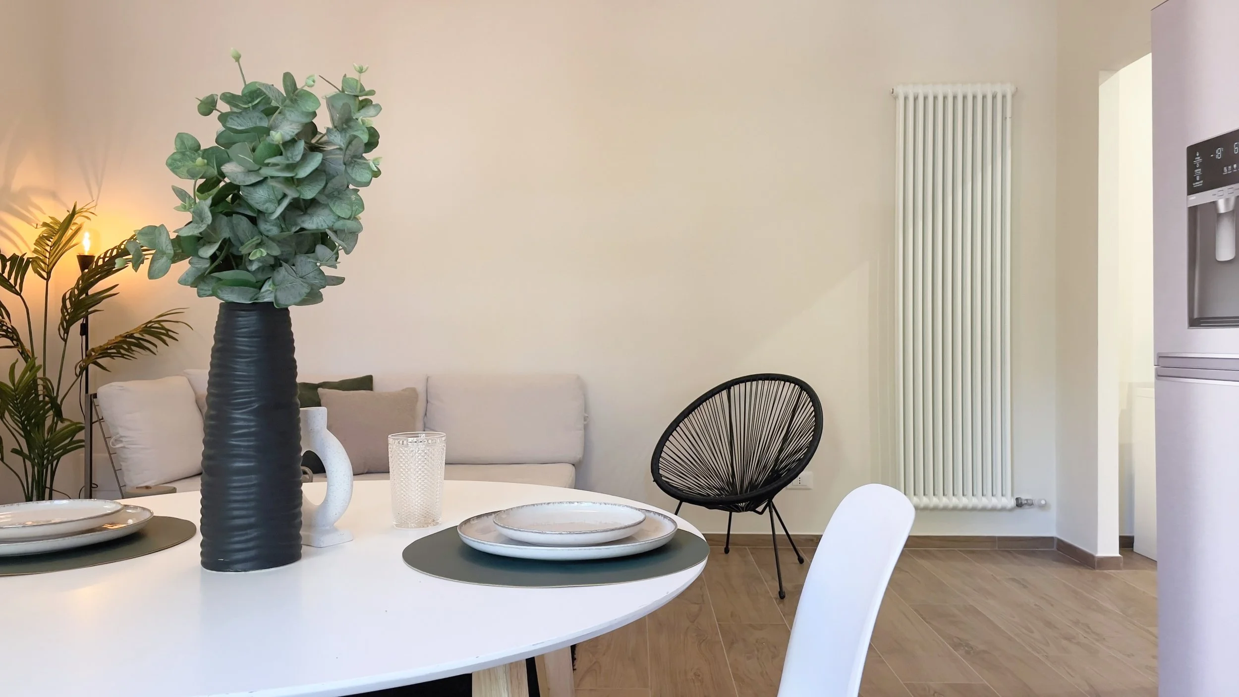 Soggiorno moderno con tavolo bianco, piatti su tovagliette verdi, vaso nero con piante, divano beige, sedia nera, lampada, e frigorifero in acciaio.