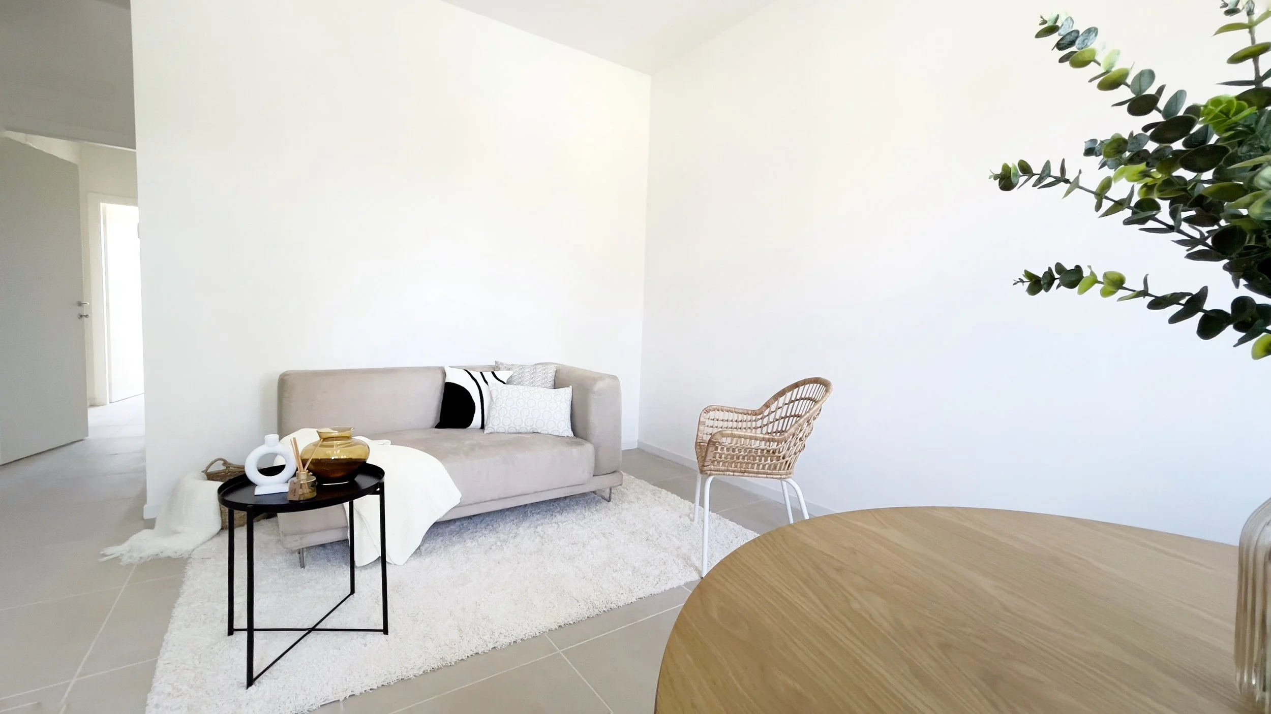 Soggiorno minimalista con divano beige, tavolino nero, sedia intrecciata e pianta decorativa. Arredamento semplice su tappeto chiaro.