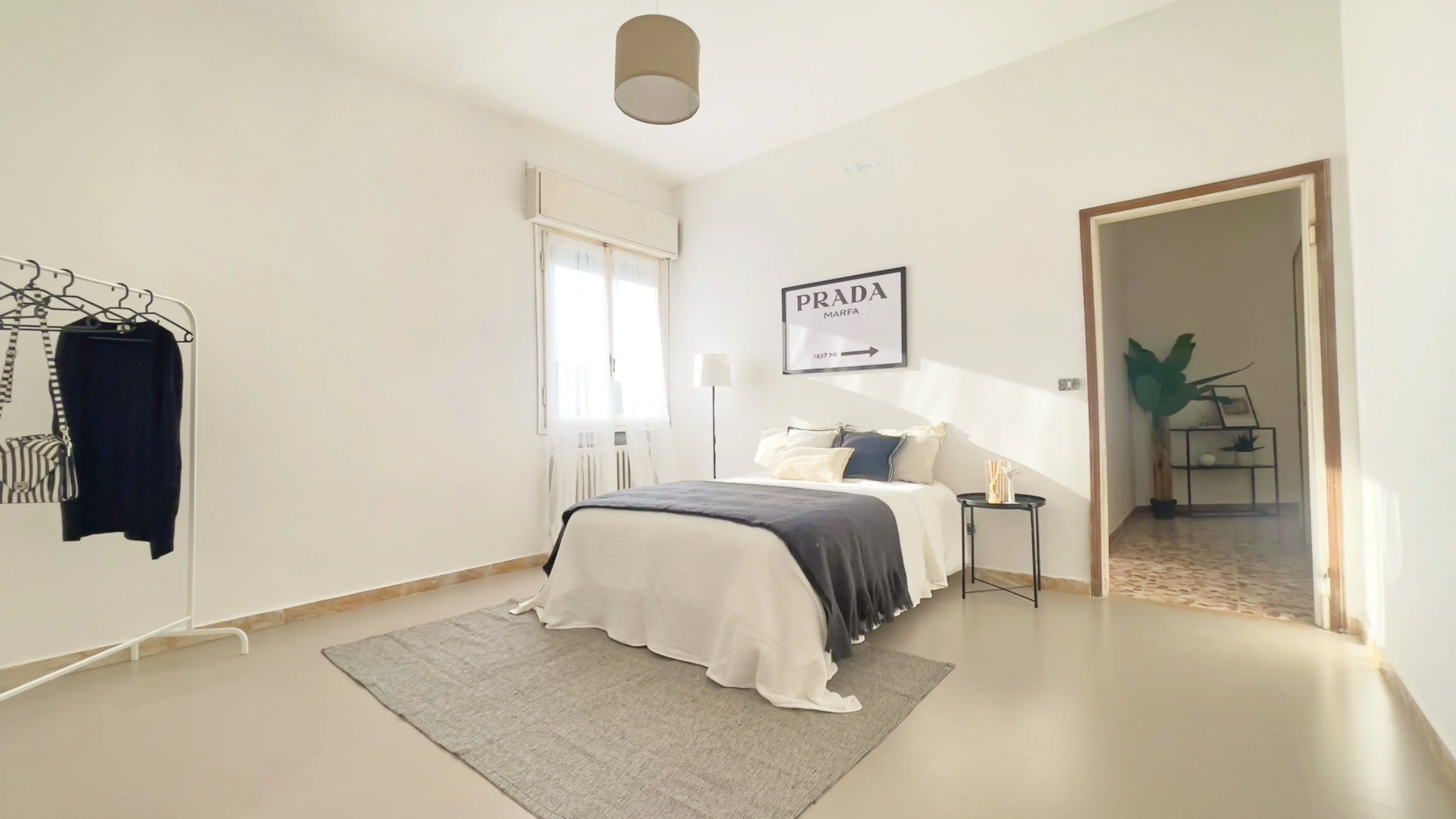 Camera da letto luminosa con letto matrimoniale coperto da coperte bianche e grigie. A destra, un comodino nero con lampada e diffusore profumato. Sulla parete, un poster. A sinistra, un appendiabiti bianco con vestiti. Dietro, una finestra illuminat