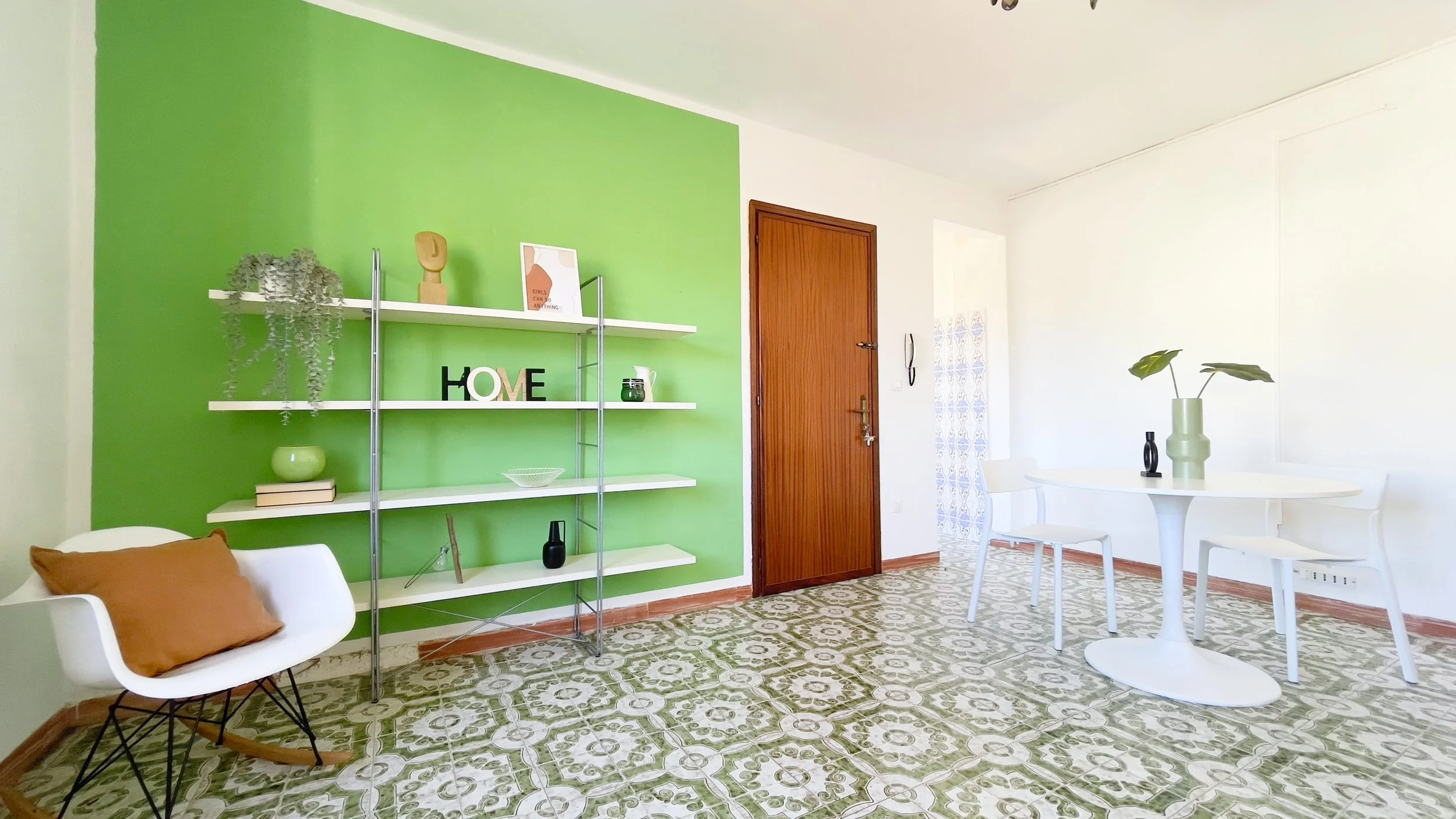 Sala luminosa con una parete verde, scaffali bianchi con decorazioni, una sedia a dondolo bianca con cuscino marrone, un tavolo bianco rotondo con sedie, e pavimento con piastrelle a motivi verdi.