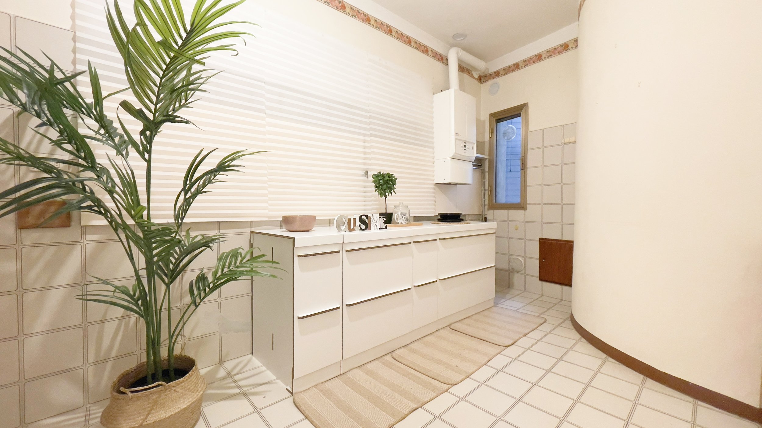 Cucina moderna con armadi bianchi, tappeti beige, pianta in vaso, finestrella e decorazioni.