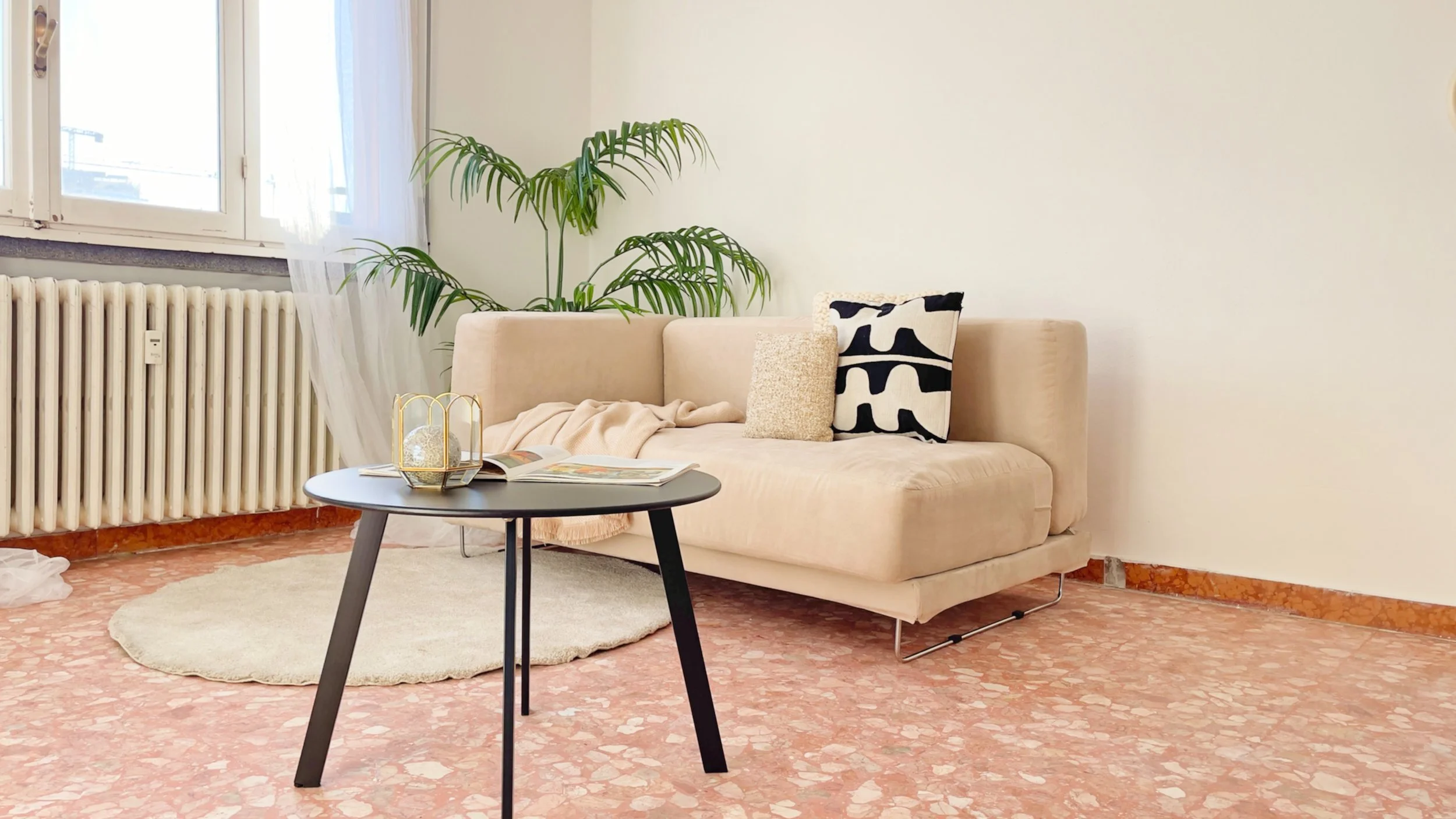Soggiorno luminoso con divano beige, cuscini decorativi, tavolino nero, pianta verde e finestra con tende leggere.