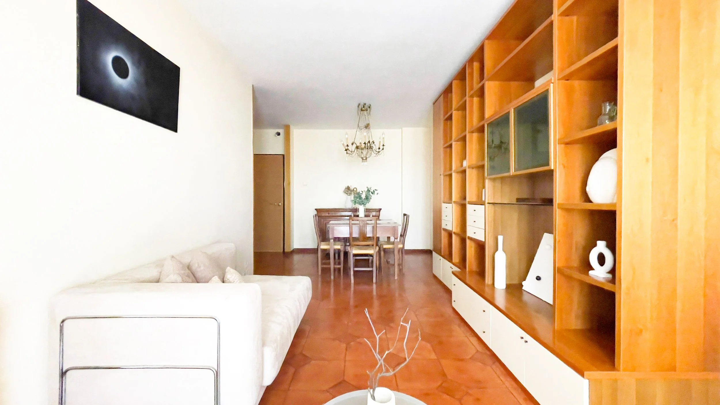 Soggiorno con divano bianco, libreria in legno, tavolo da pranzo con sedie, lampadario classico e quadro appeso.