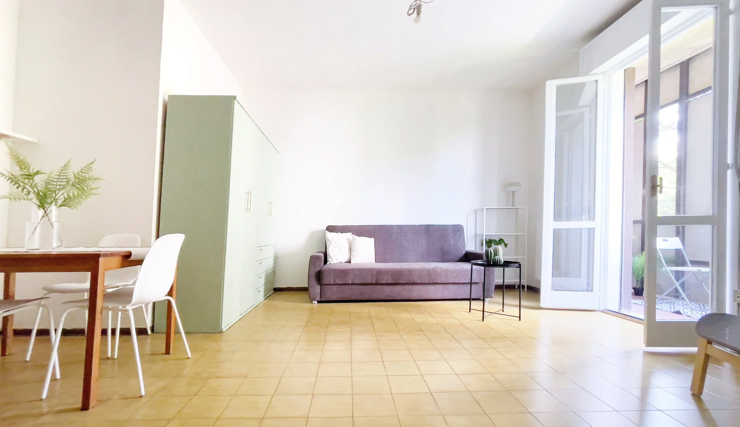 Interno di un soggiorno moderno e luminoso con un divano grigio, tavolo da pranzo con sedie bianche, armadio verde chiaro, piantana e tavolino con pianta. Ampia finestra aperta a destra.
