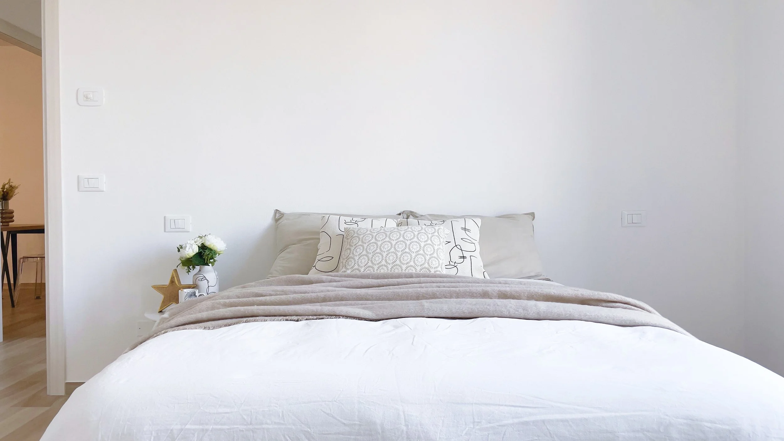 Camera da letto minimalista con letto matrimoniale, cuscini decorativi, coperta grigia e parete bianca. Tavolino con vaso di fiori bianchi e stella dorata.