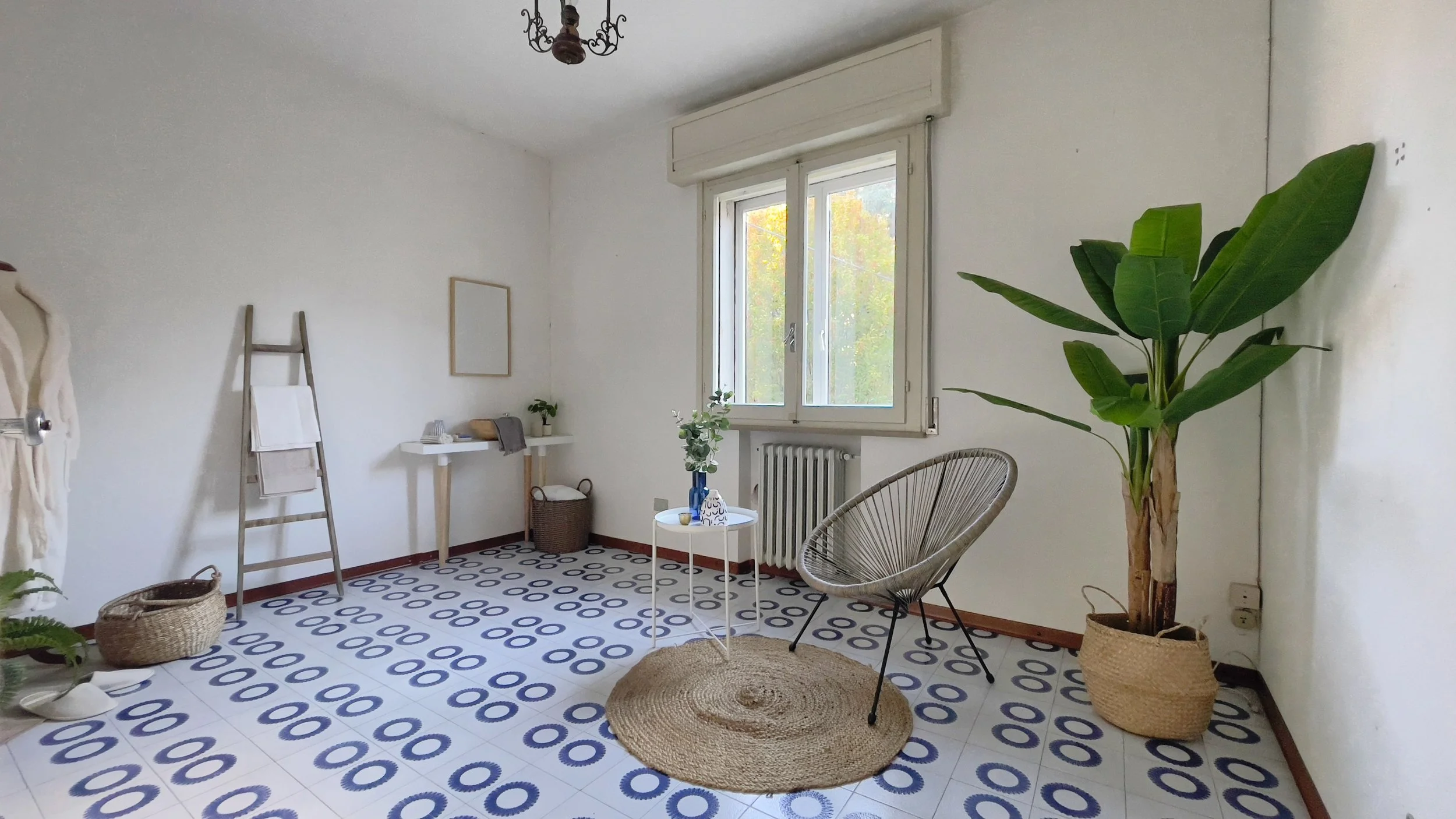 Stanza luminosa con pavimento a motivi blu, sedia moderna, tavolino rotondo bianco, pianta verde in un vaso di vimini, scala in legno con asciugamani appesi, e un manichino con accappatoio.